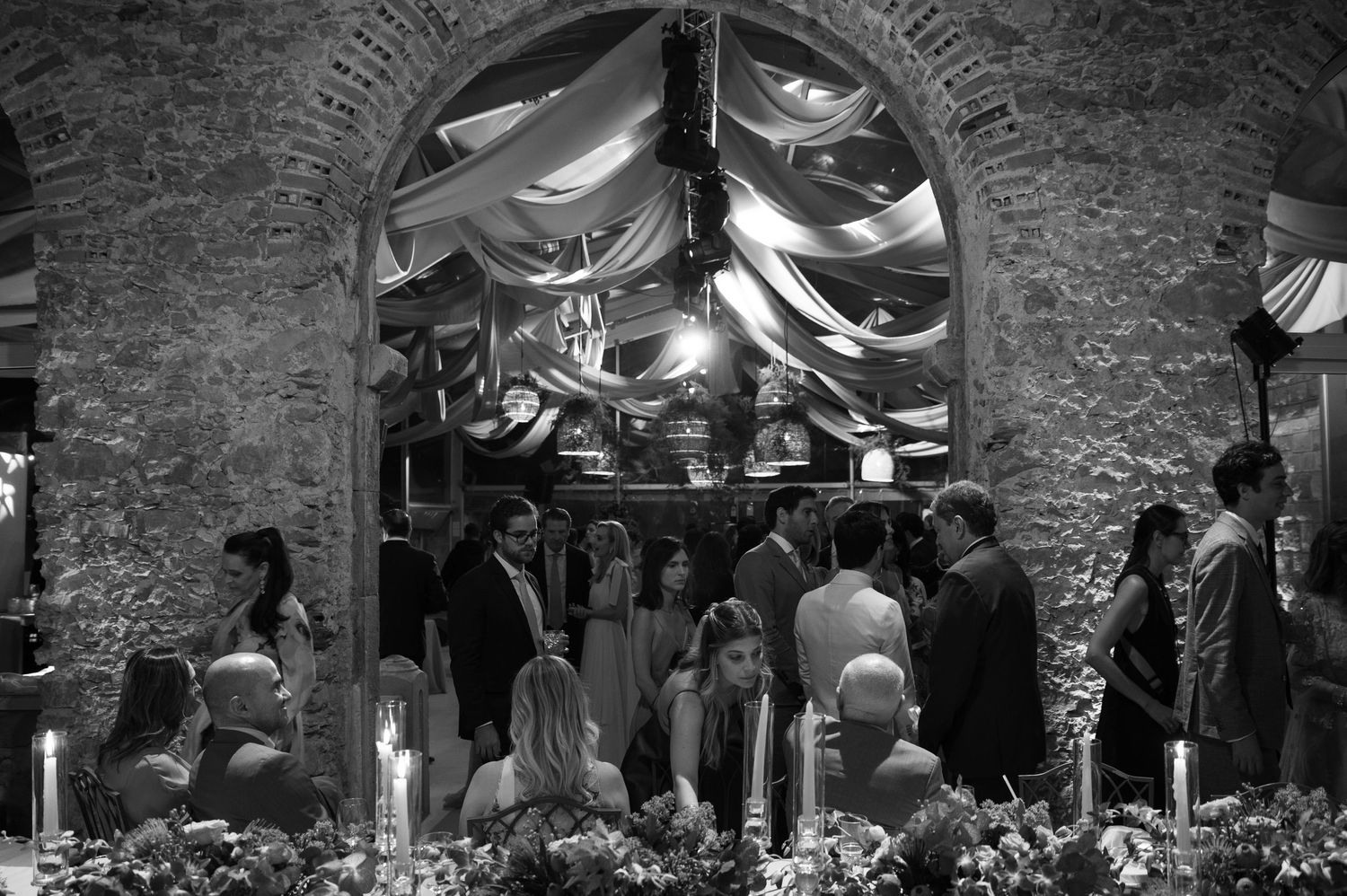Black and white photo of wedding reception in historic arched stone cellar with hanging fabric decor.