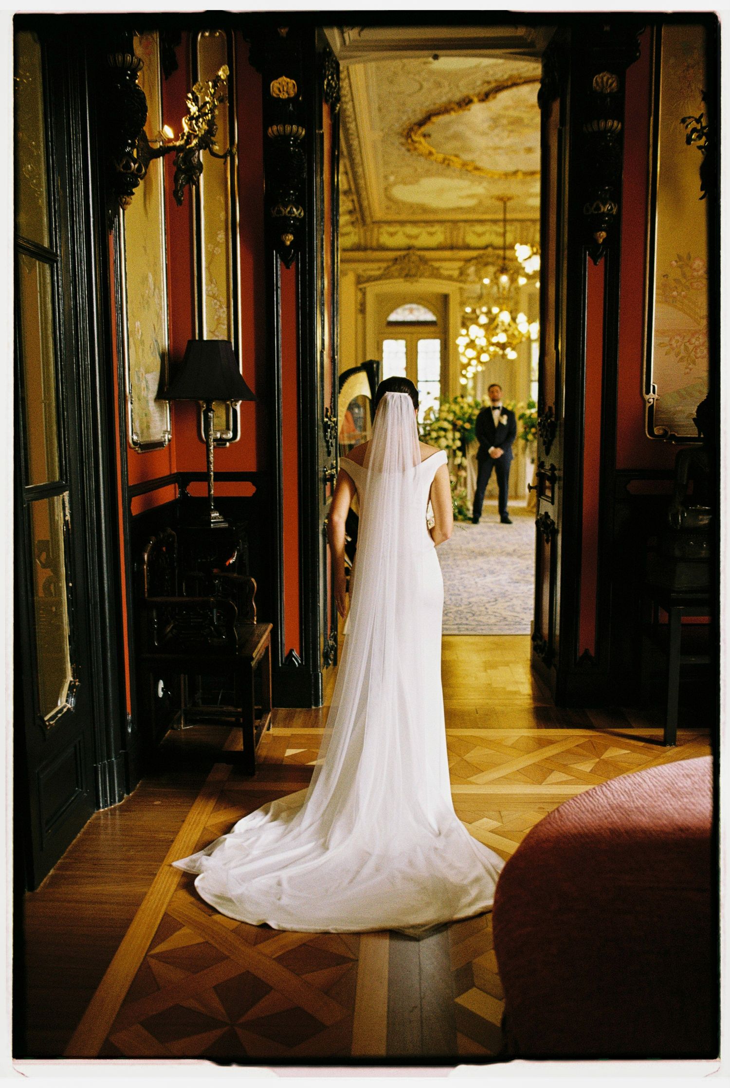 Bride in white gown and veil stands in ornate doorway facing groom in elegant venue.
