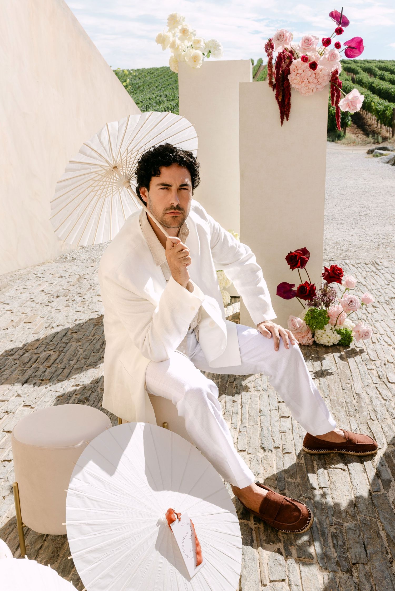 Man in white linen suit holding parasol surrounded by floral arrangements at outdoor vineyard wedding venue.