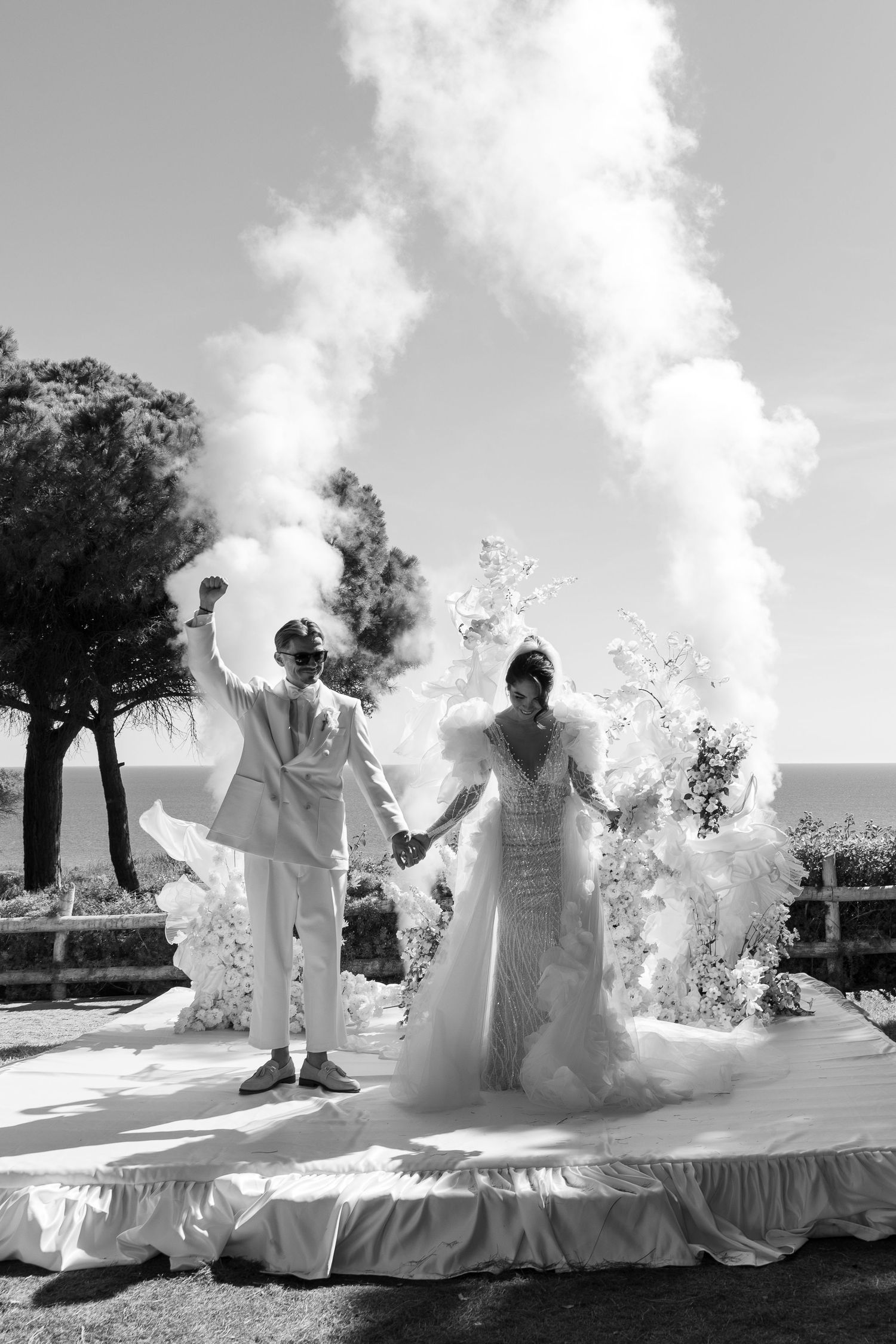 Wedding couple in white outfits creating dramatic smoke effects during outdoor photoshoot.