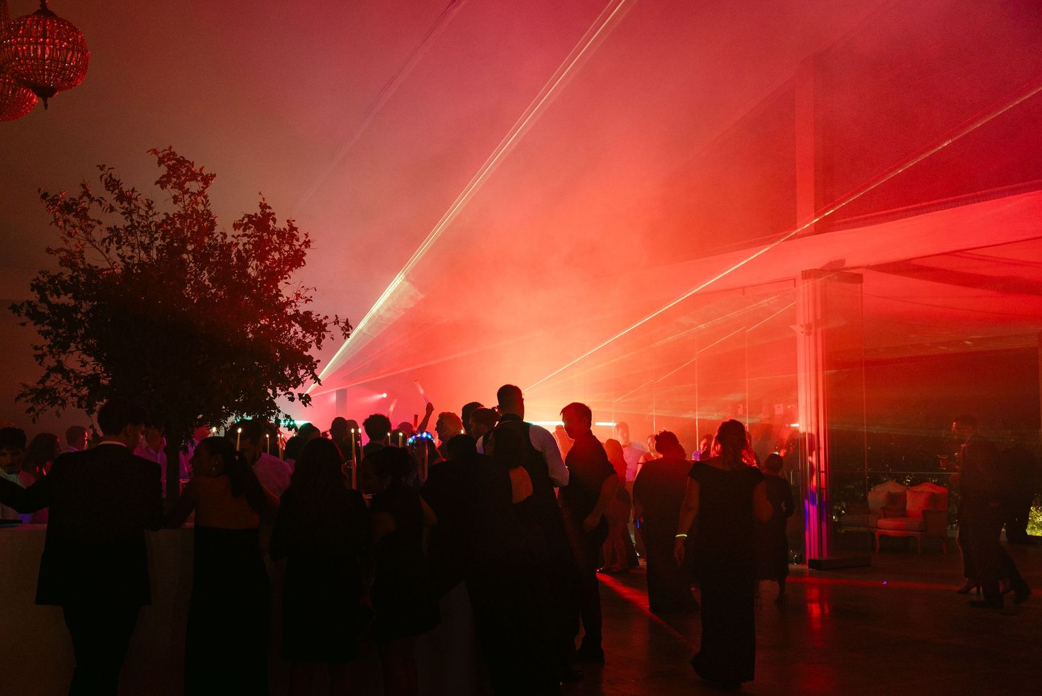 A crowd gathers under red lighting at an outdoor nighttime event with tent structures.