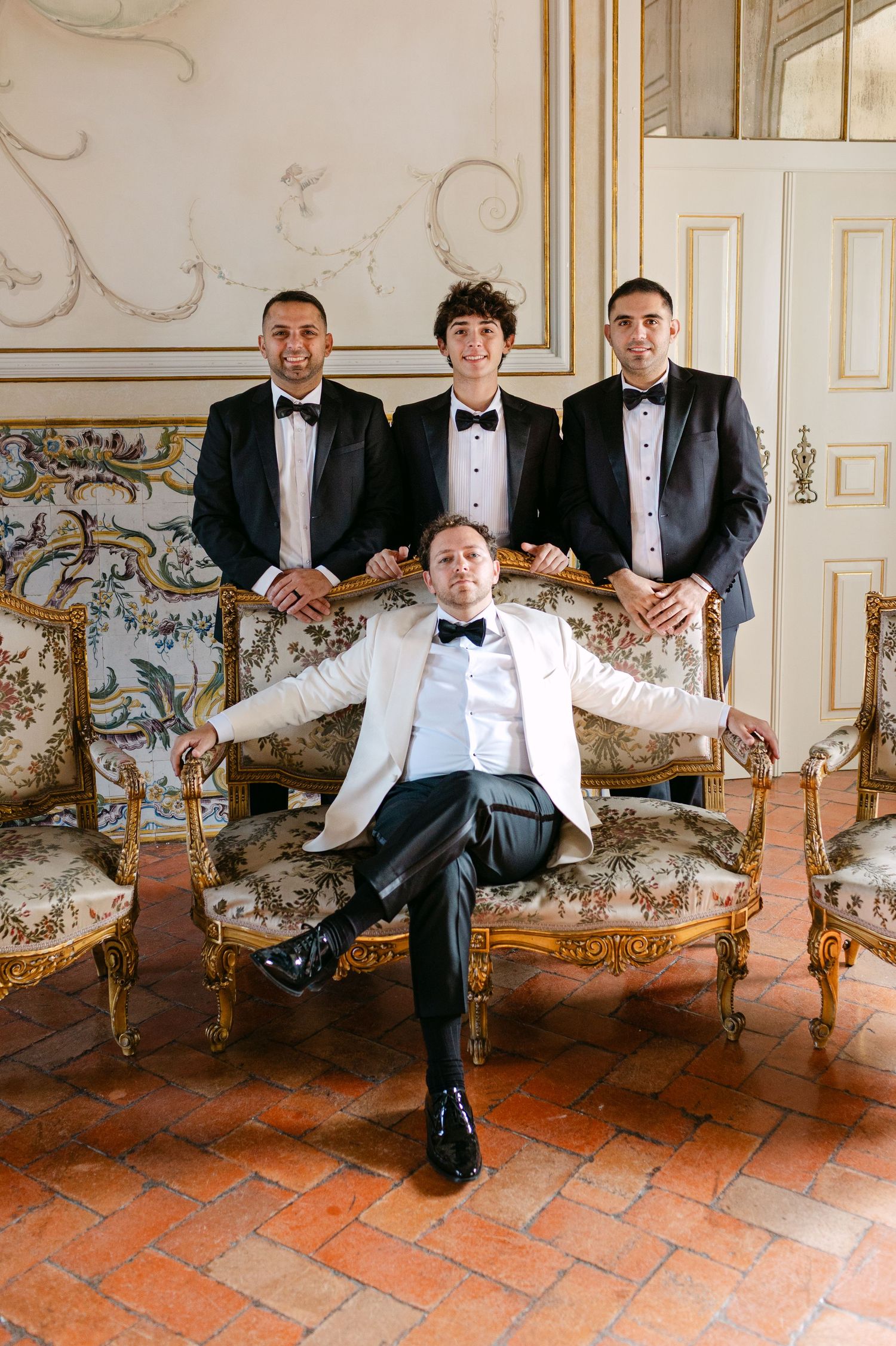 Wedding party relaxes on antique furniture in an ornately decorated room with painted walls.