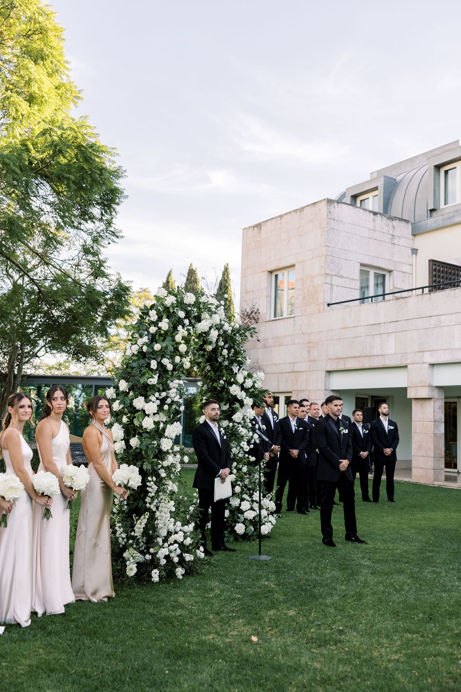 Wedding party members in formal attire gather on green lawn with modern building backdrop.
