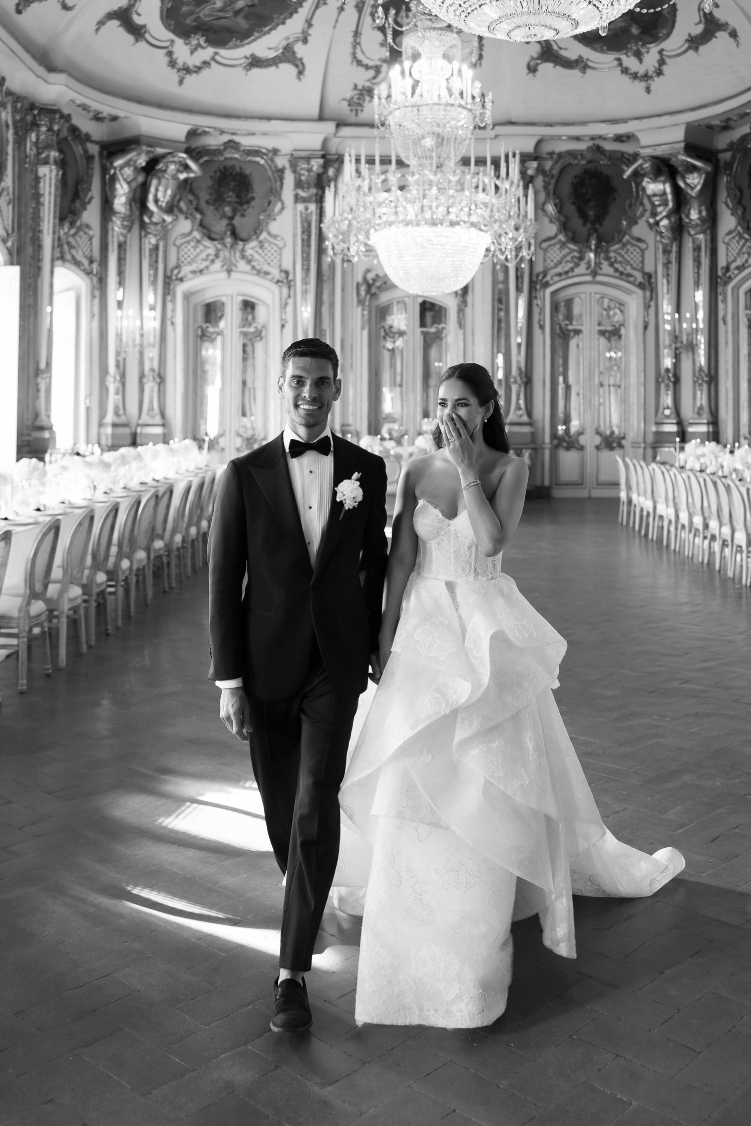 Elegant couple in formal attire walking through ornate palace ballroom.