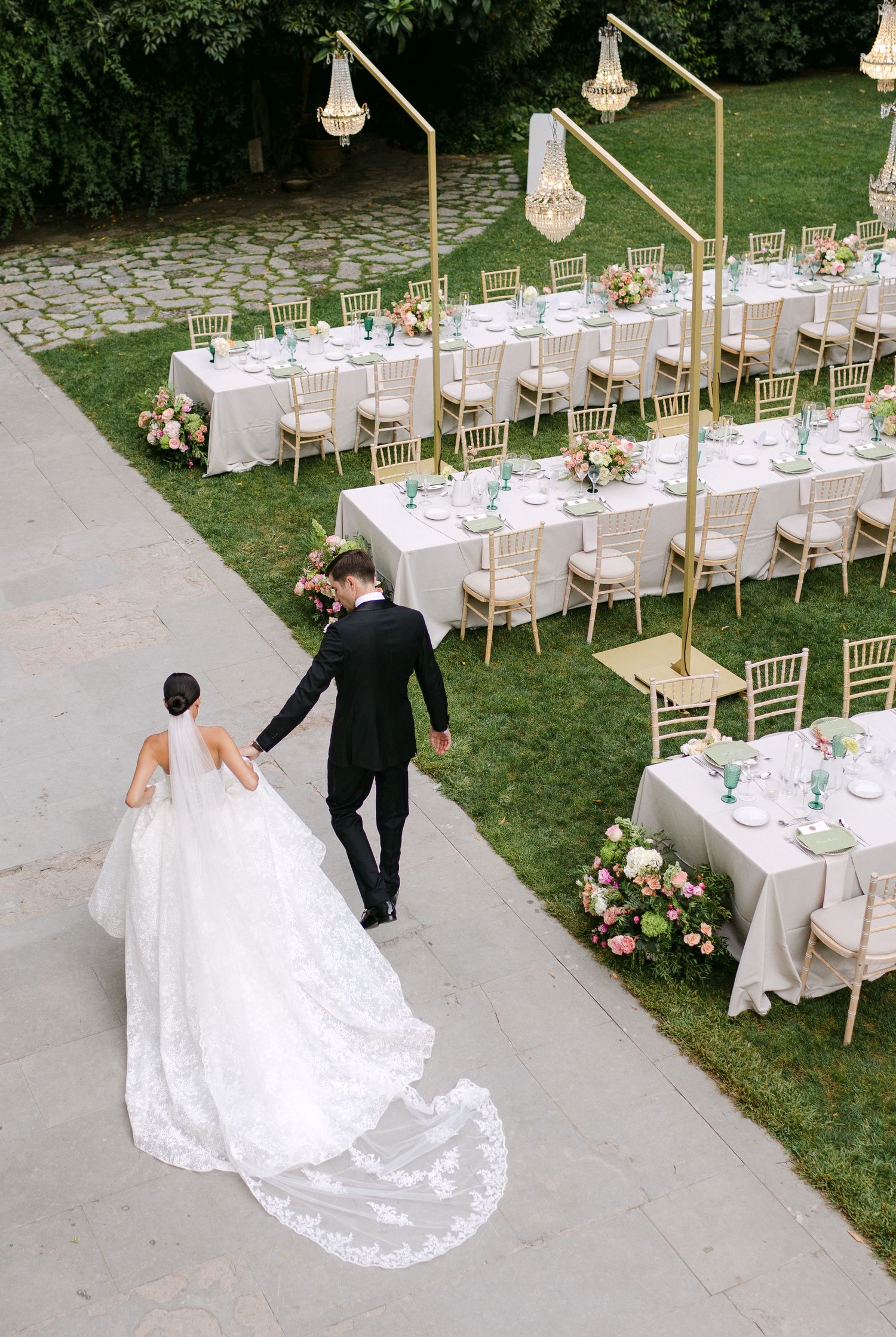 Elegant outdoor wedding reception setup with white tables, gold chairs, and hanging crystal chandeliers on a garden lawn.