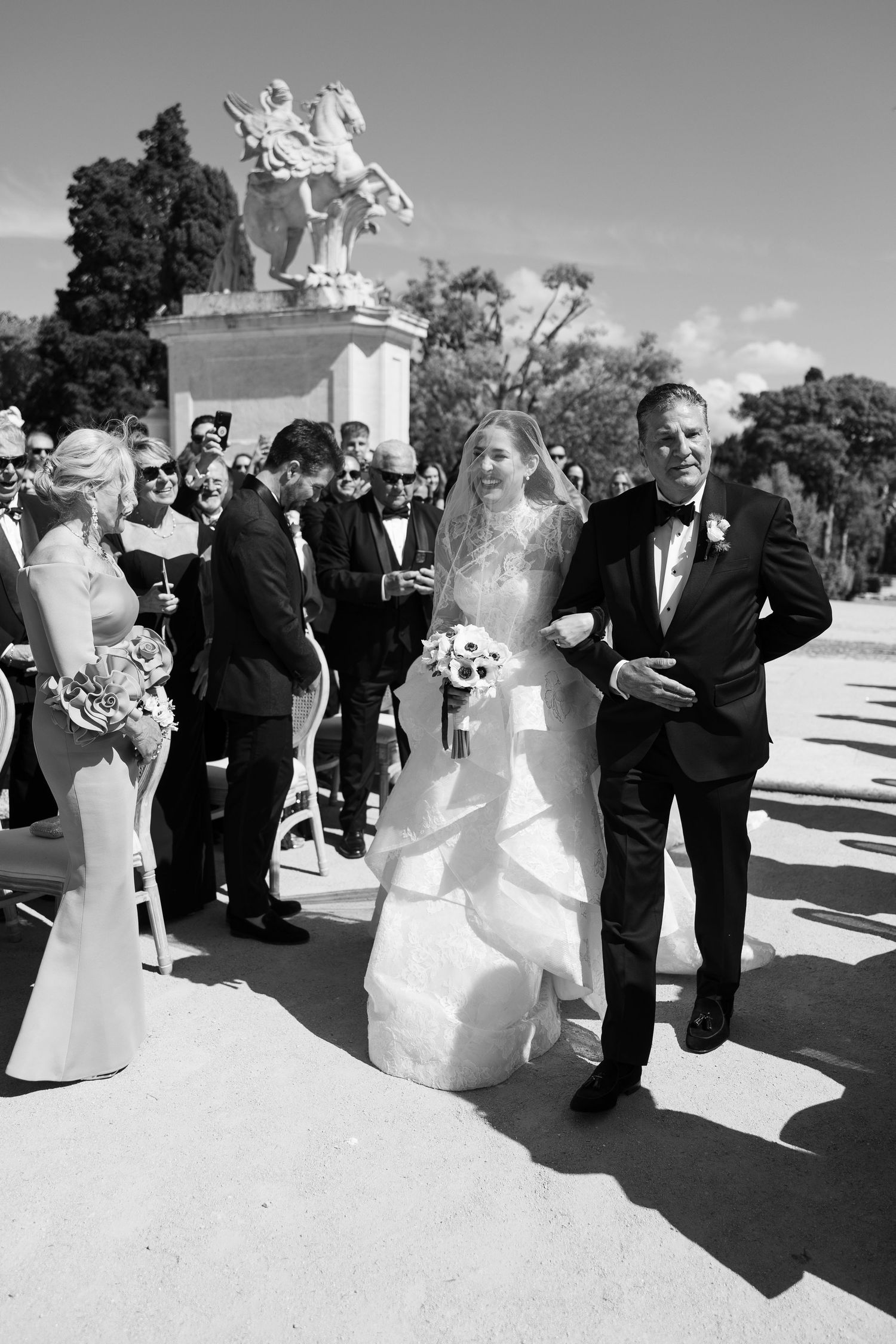 Bride walks down the aisle with escort at outdoor wedding ceremony near classical statue.