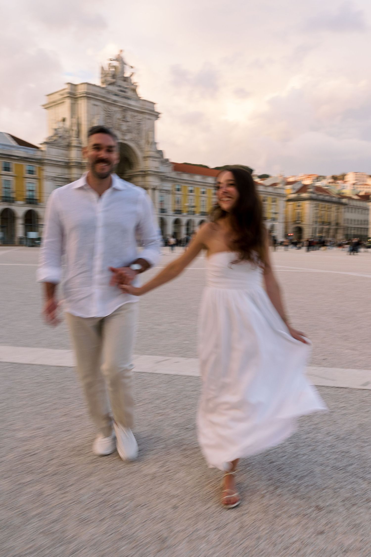A joyful couple in white outfits holds hands while walking through Lisbon's Praca do Comercio at sunset.