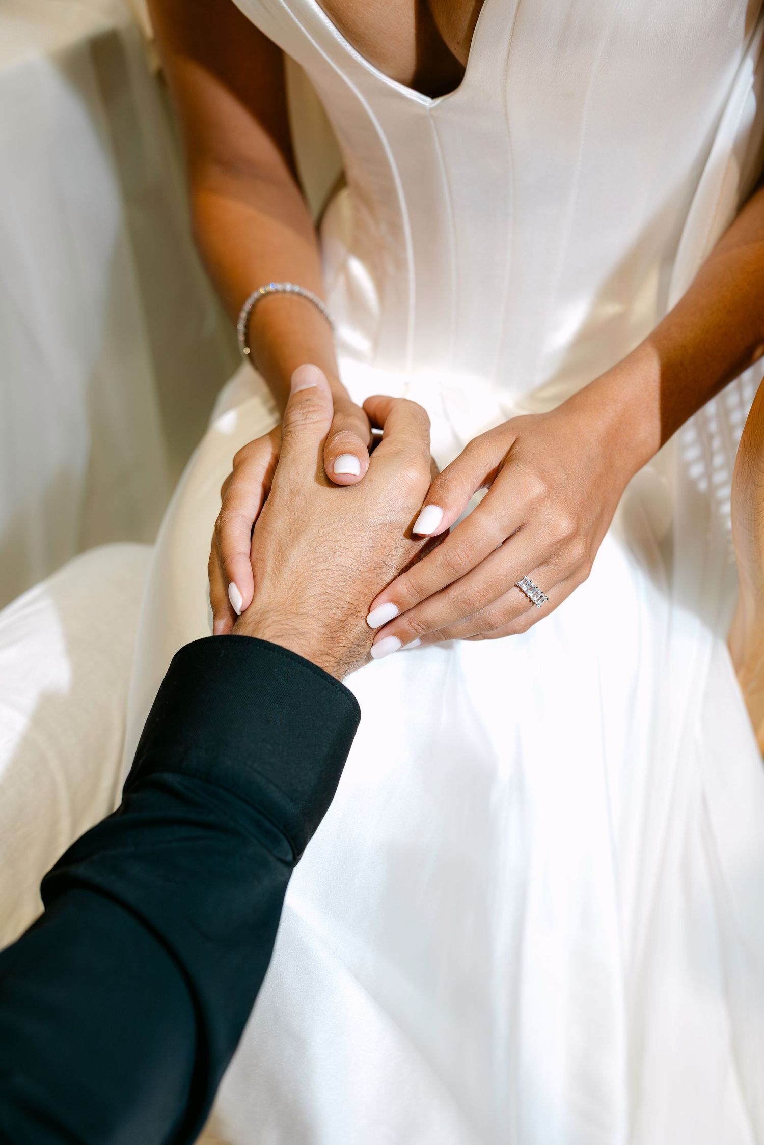 Close-up of bride and groom holding hands, bride wearing diamond ring and pearl bracelet in white gown.