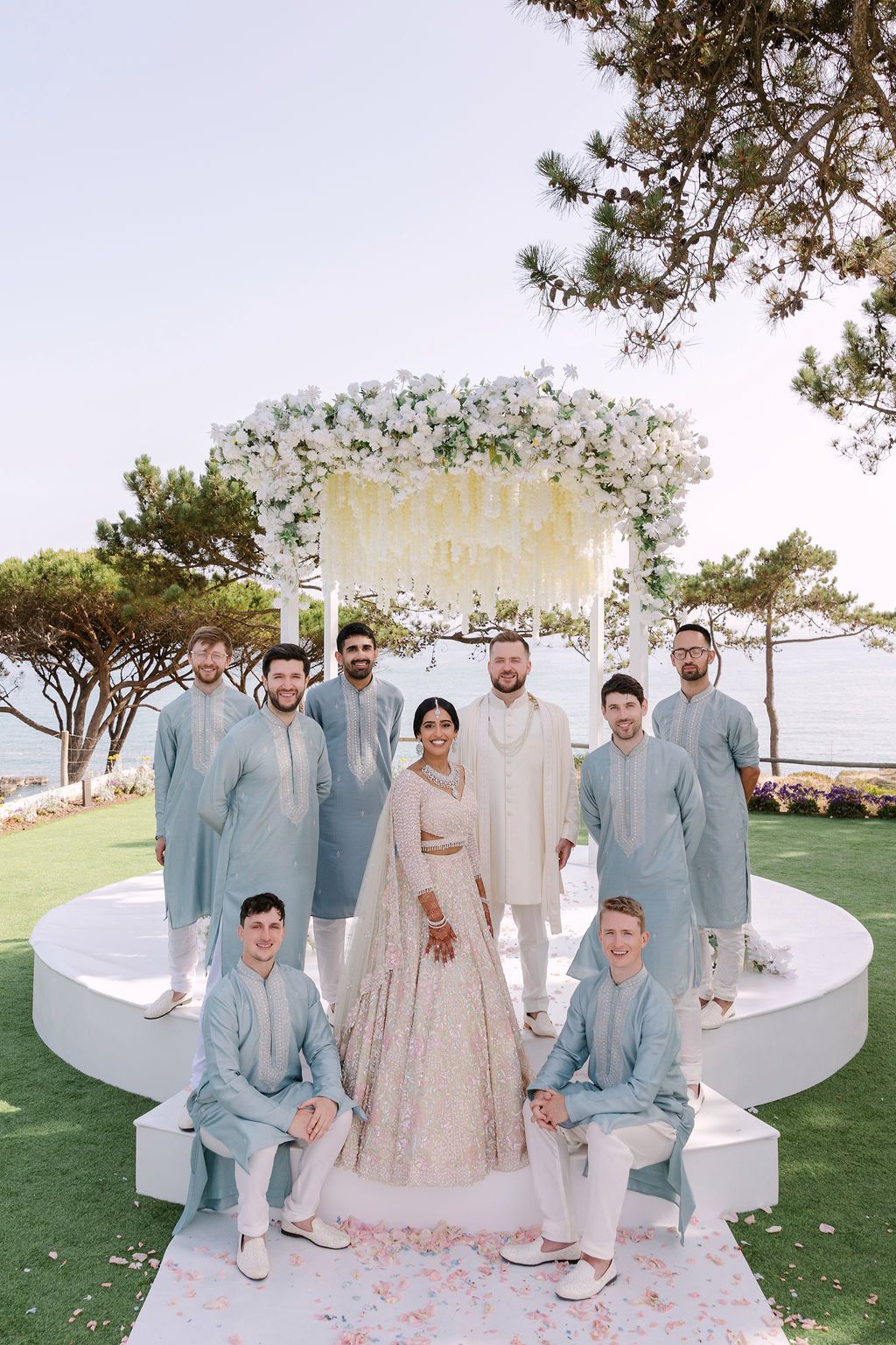Wedding party in light blue suits and cream dress pose together on white circular platform with ocean view.
