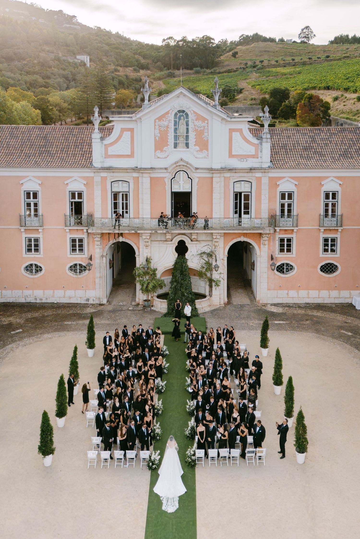 Aerial view of a wedding ceremony setup in front of a pink historic mansion with symmetrical architecture and cypress trees.
