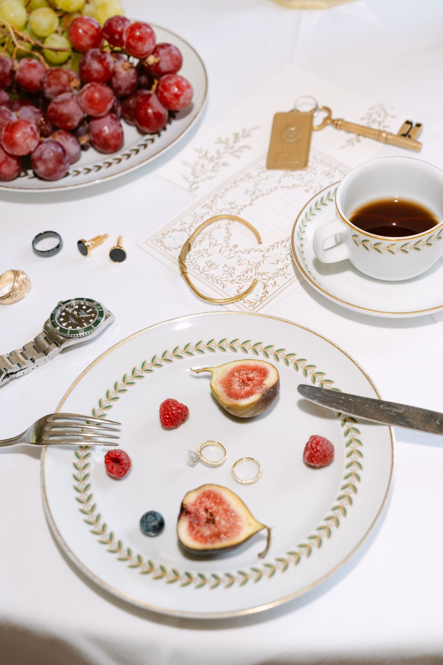 Elegant place setting with gold-rimmed plates, fresh figs, and grapes on white tablecloth.