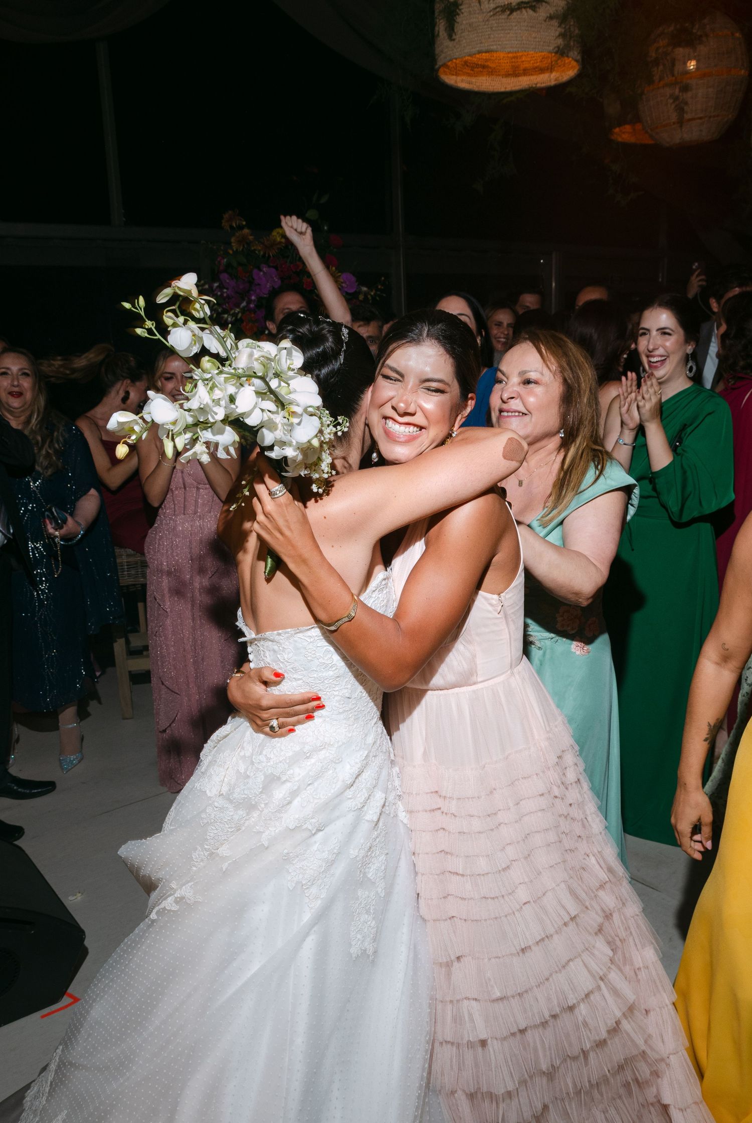Wedding guests in white and pastel dresses dance together at an indoor reception.