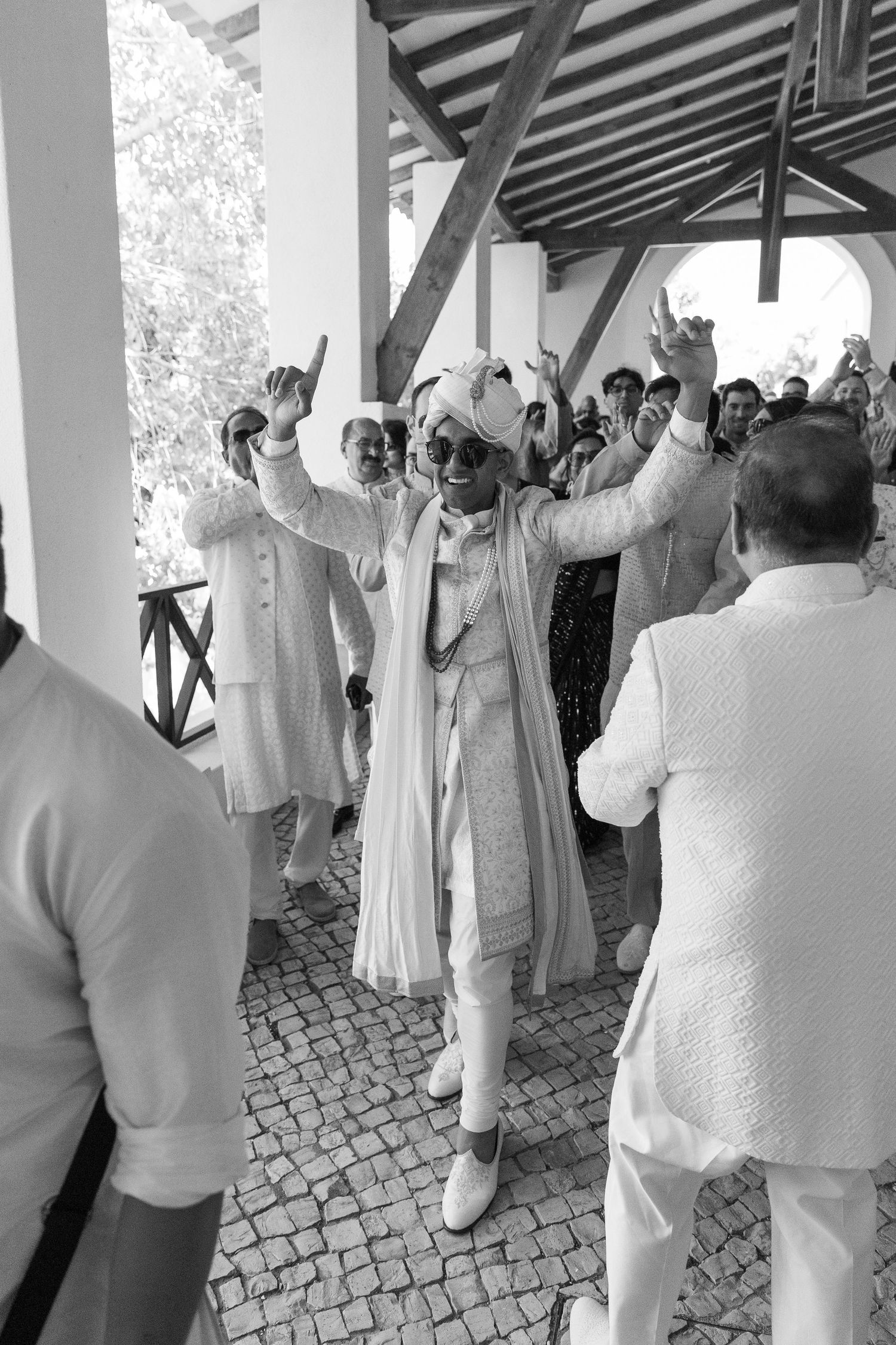 Wedding party dressed in white performs celebratory dance moves under covered outdoor venue.