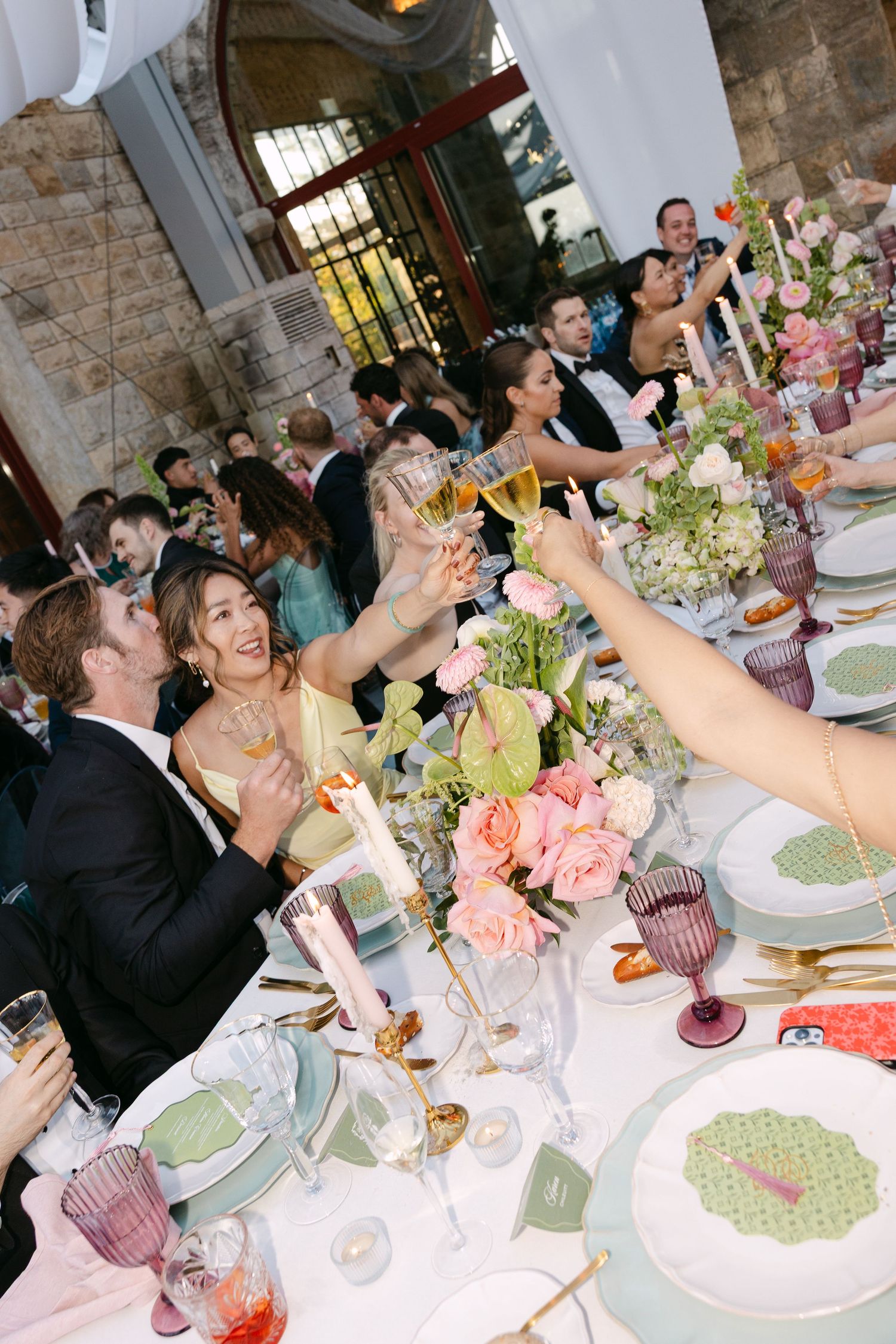 Wedding guests raise their glasses in a toast at an elegant reception with pink floral centerpieces and vintage decor.