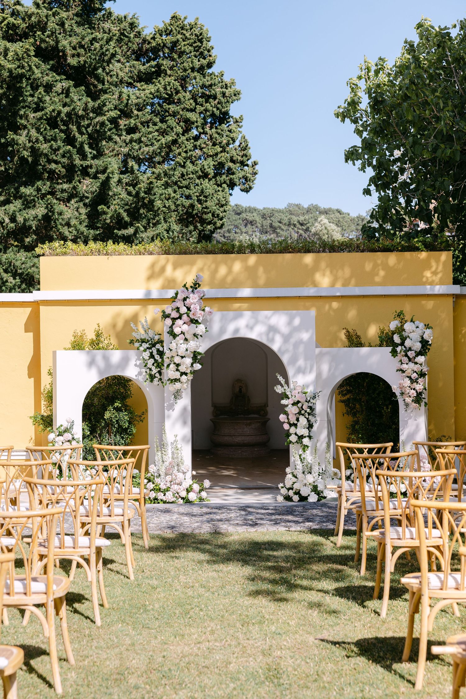 Outdoor wedding ceremony setup with wooden chairs facing white floral arch and yellow building.