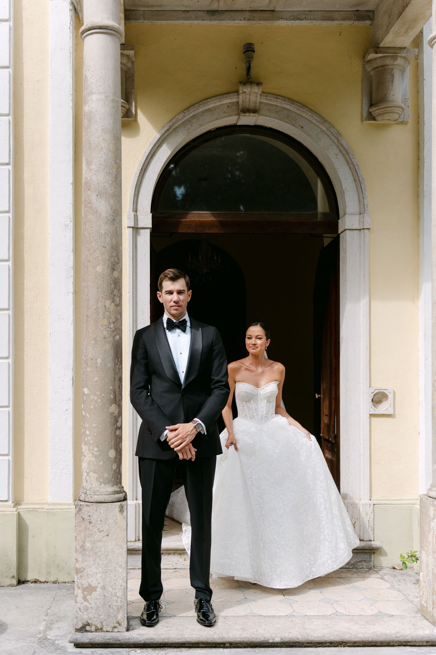 Elegant couple in wedding attire standing under arched doorway of classic building.