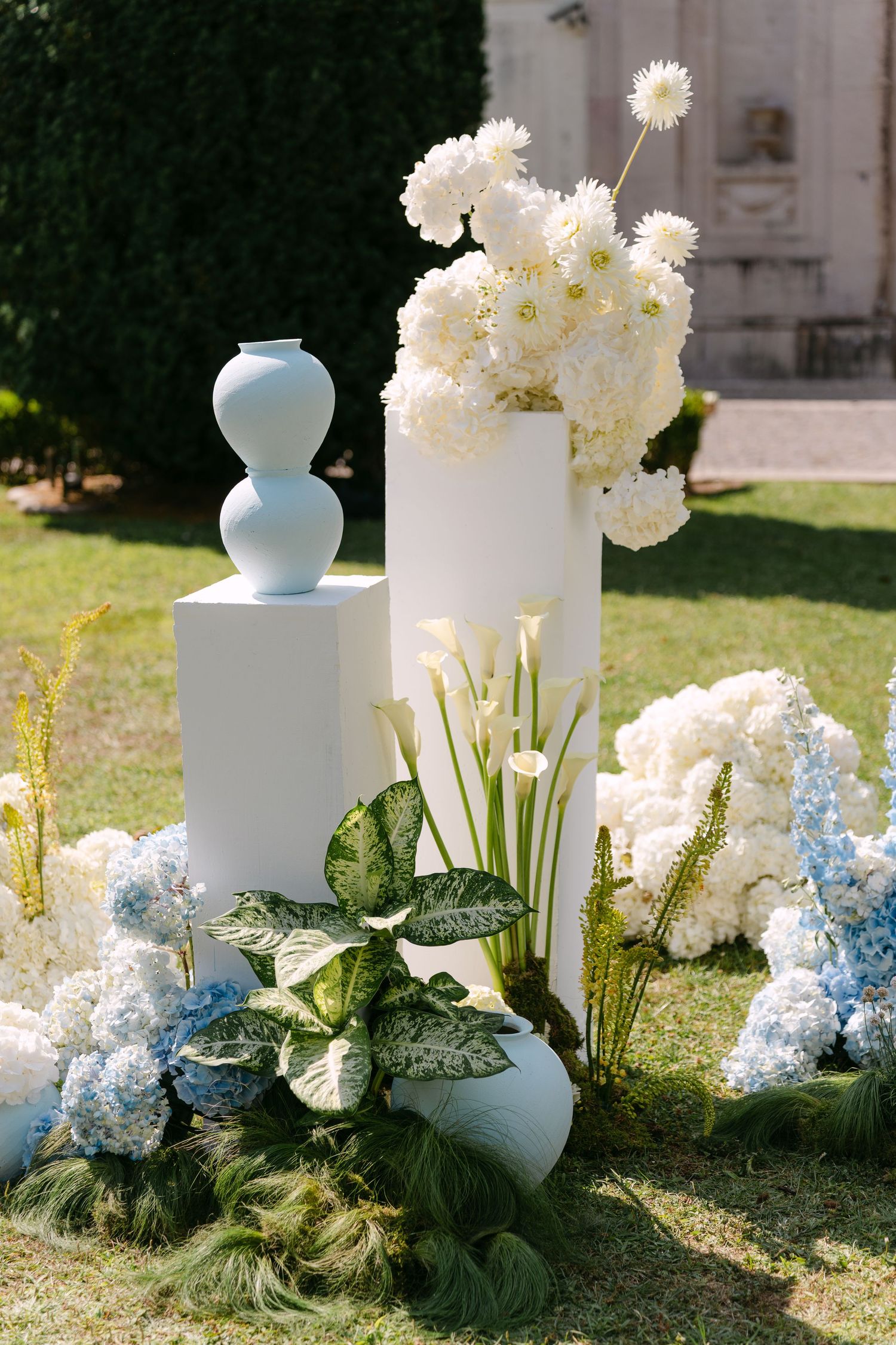 White floral arrangements with blue hydrangeas displayed on pedestals against green lawn.