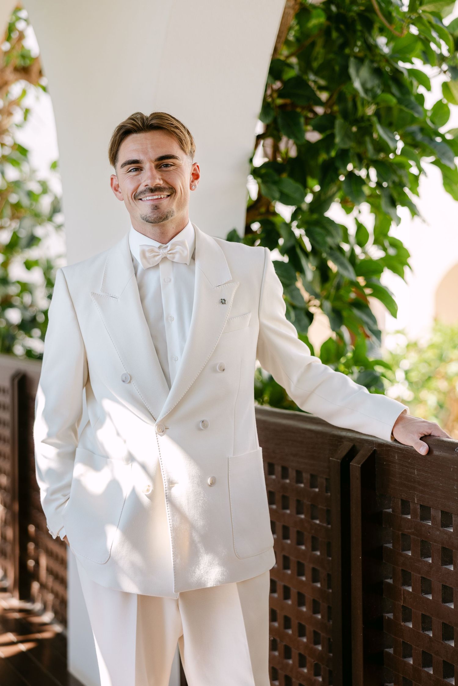 A formal white double-breasted suit with bow tie stands in an elegant outdoor setting.