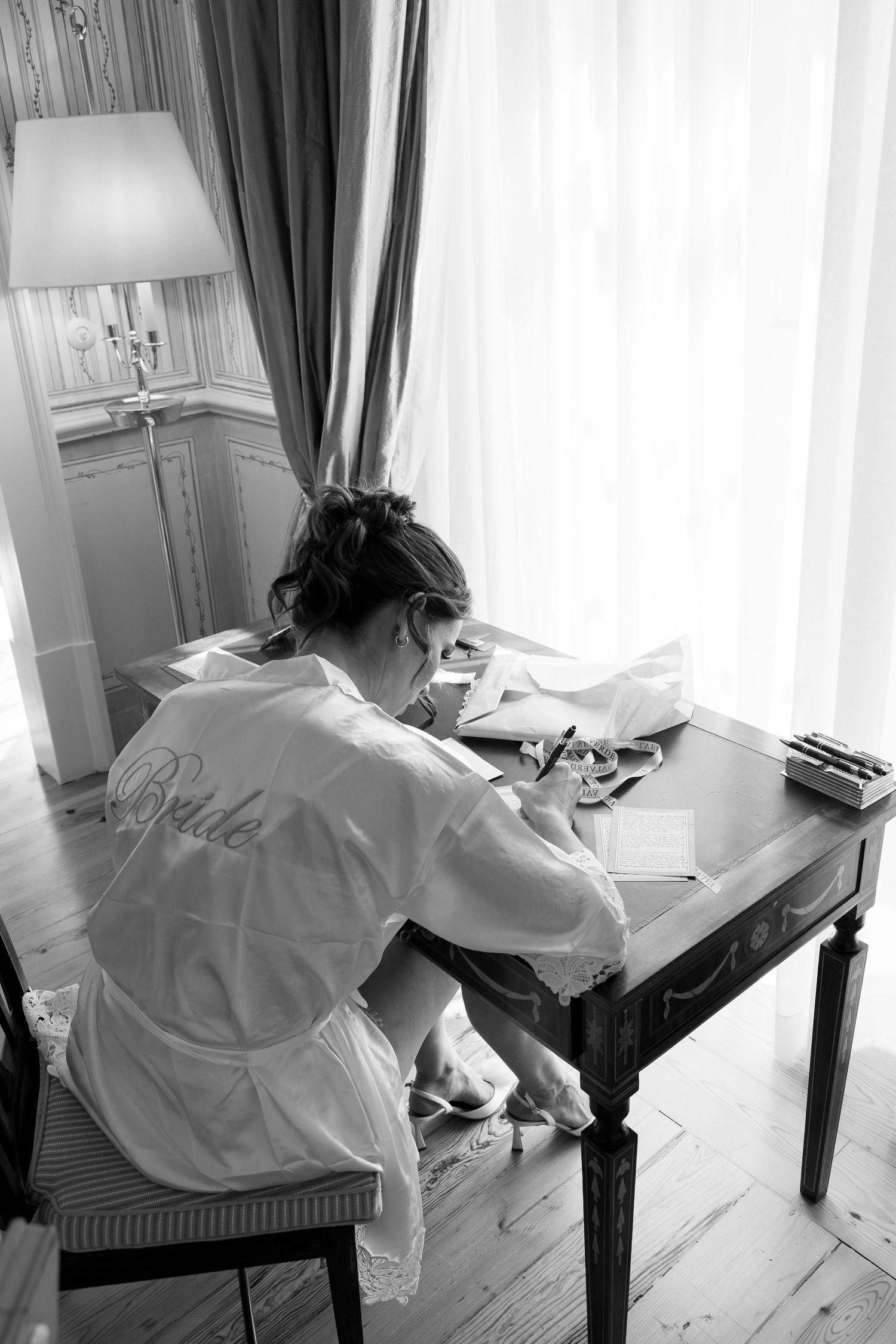 Person in white robe writing at a desk near window in black and white photography.