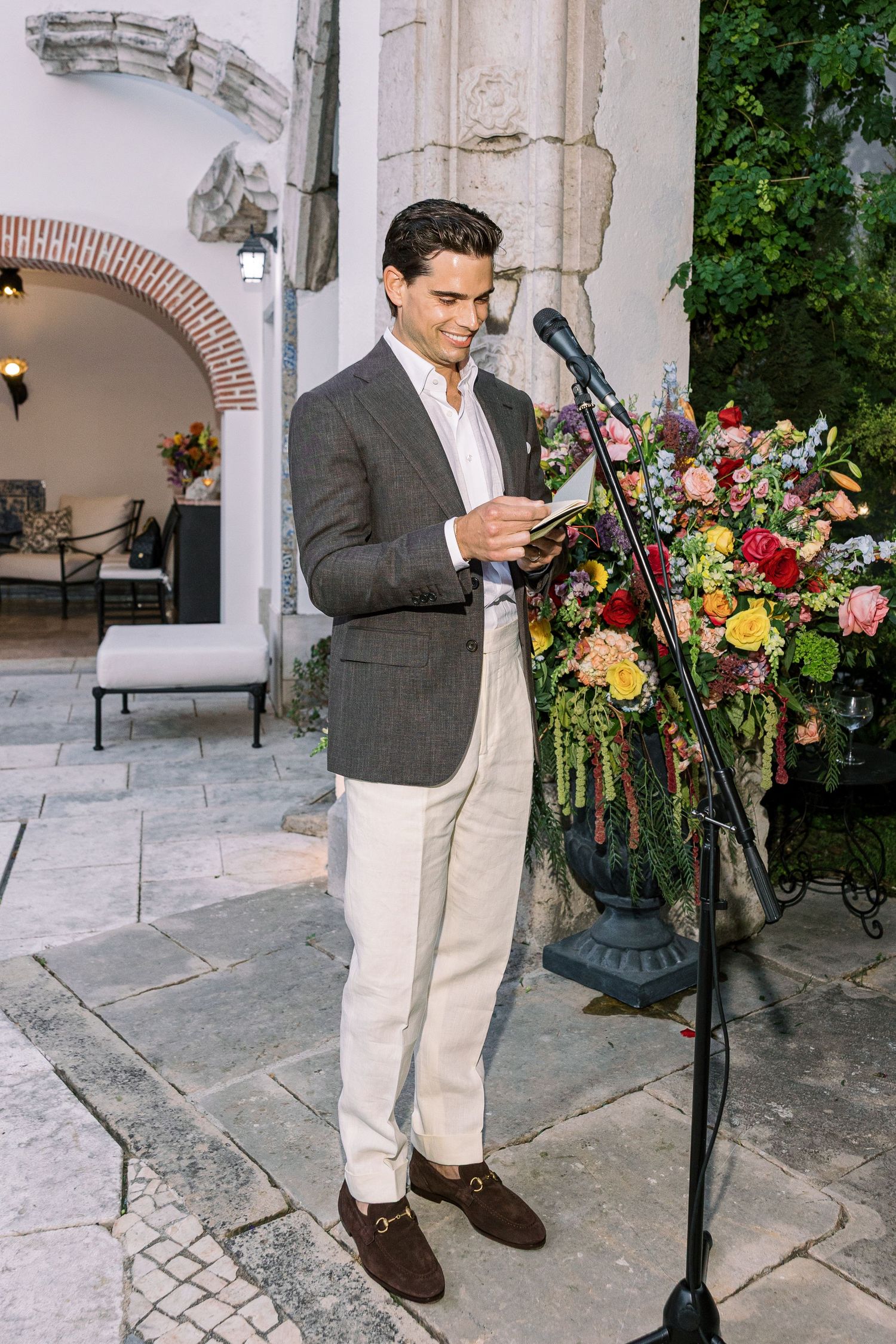 Elegant man in gray blazer and cream trousers reads at a microphone beside colorful floral arrangement at outdoor event.