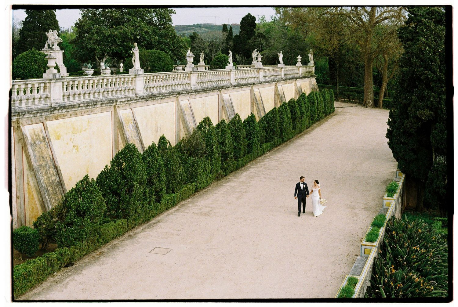 Bride and groom walking along elegant garden path with ornate balustrade wall and manicured hedges.