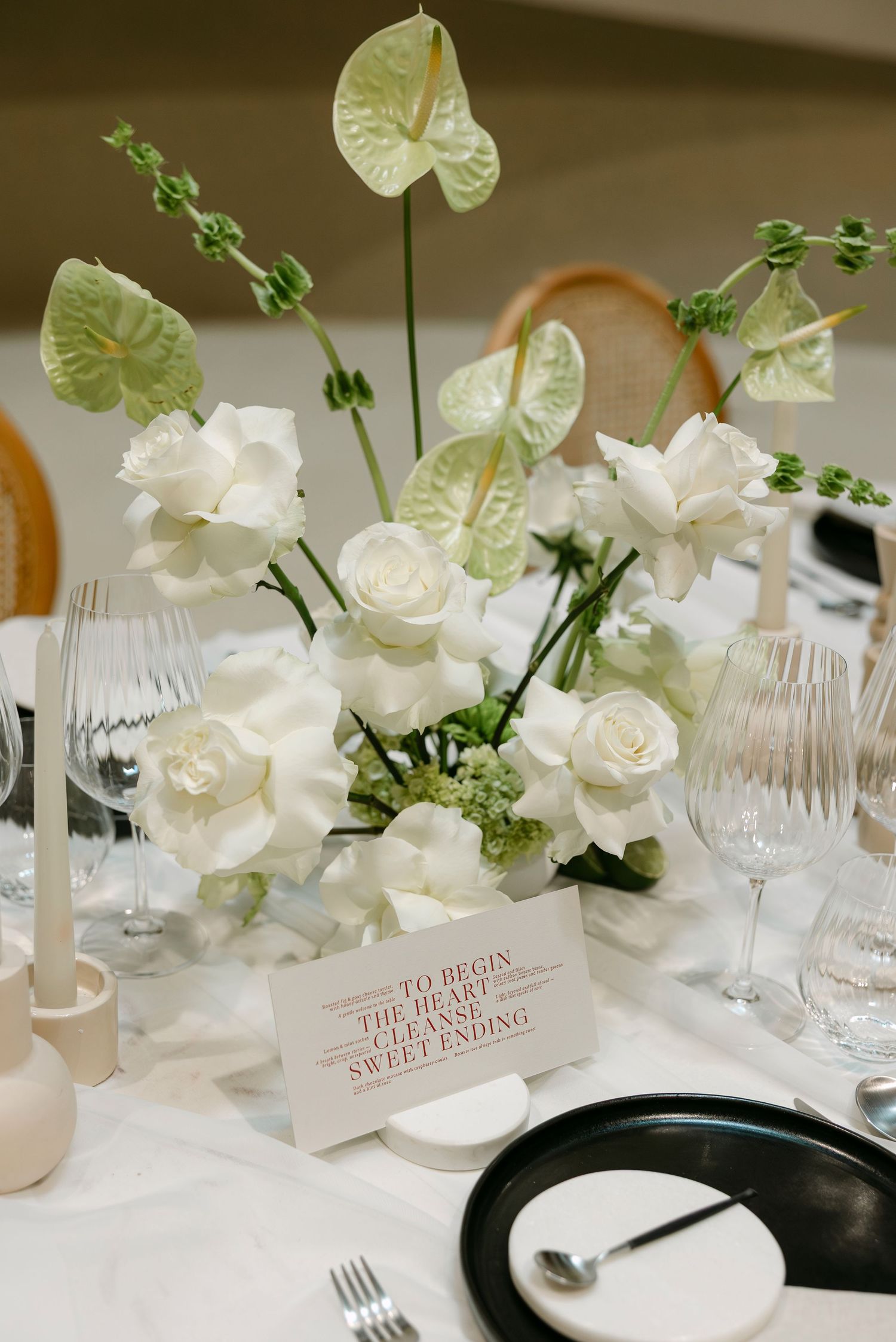 Elegant white floral centerpiece with anthuriums and roses beside a menu card and crystal glassware on a white tablecloth.