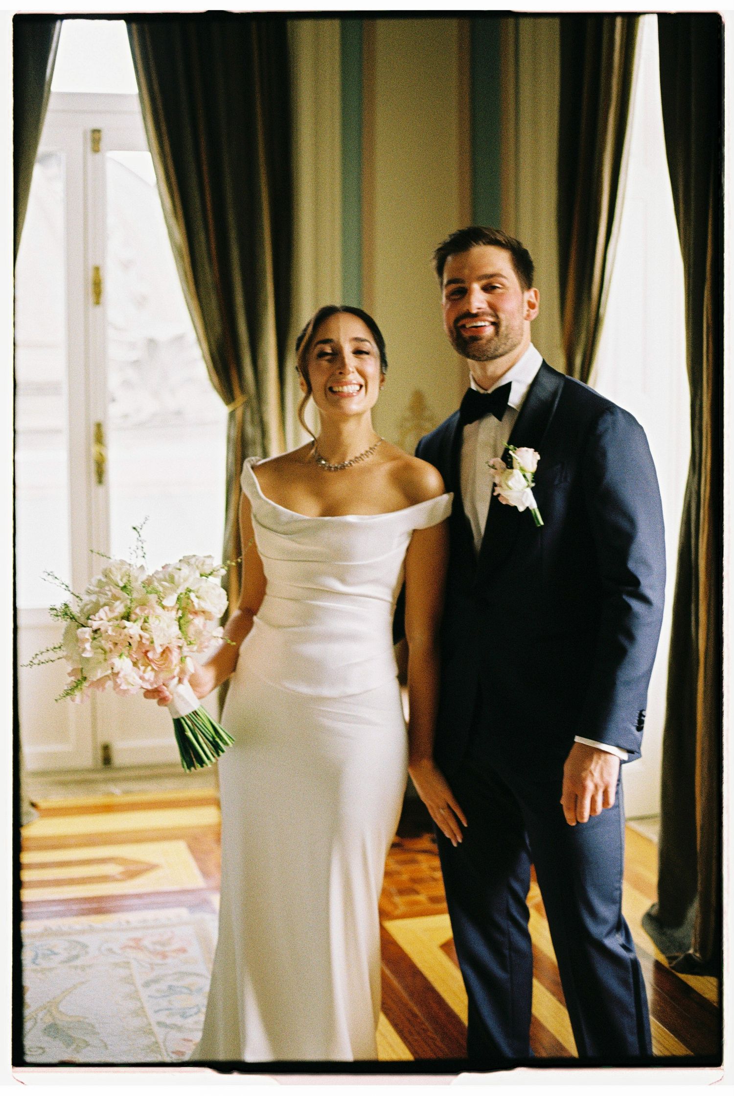 Bride in white off-shoulder gown holding pink bouquet stands with groom in navy tuxedo at elegant wedding venue.