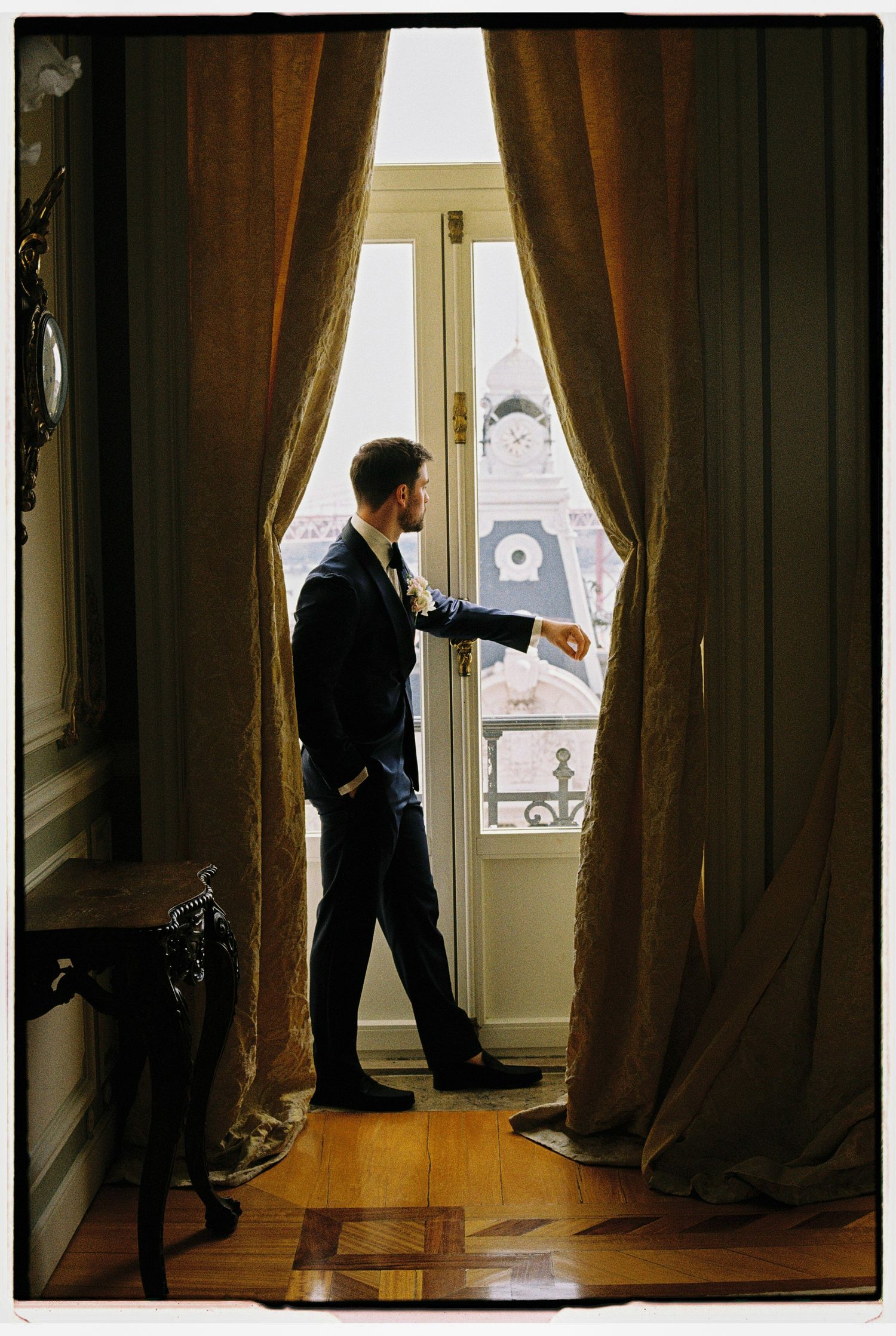 Dapper groom in a navy suit standing by a grand window with flowing curtains checking his watch.