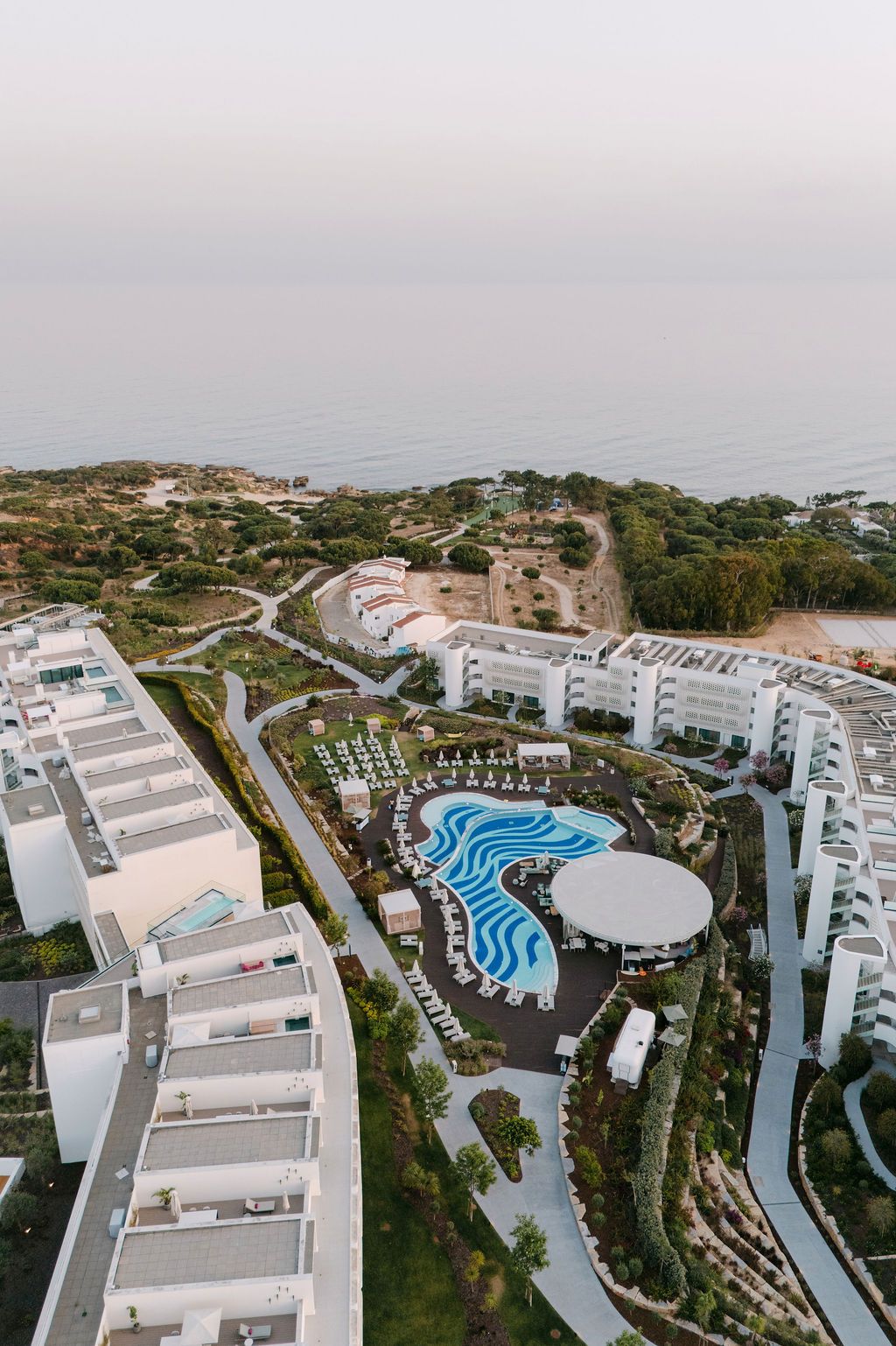 Aerial view of a luxury resort complex with white buildings and curved swimming pools overlooking the coastline.