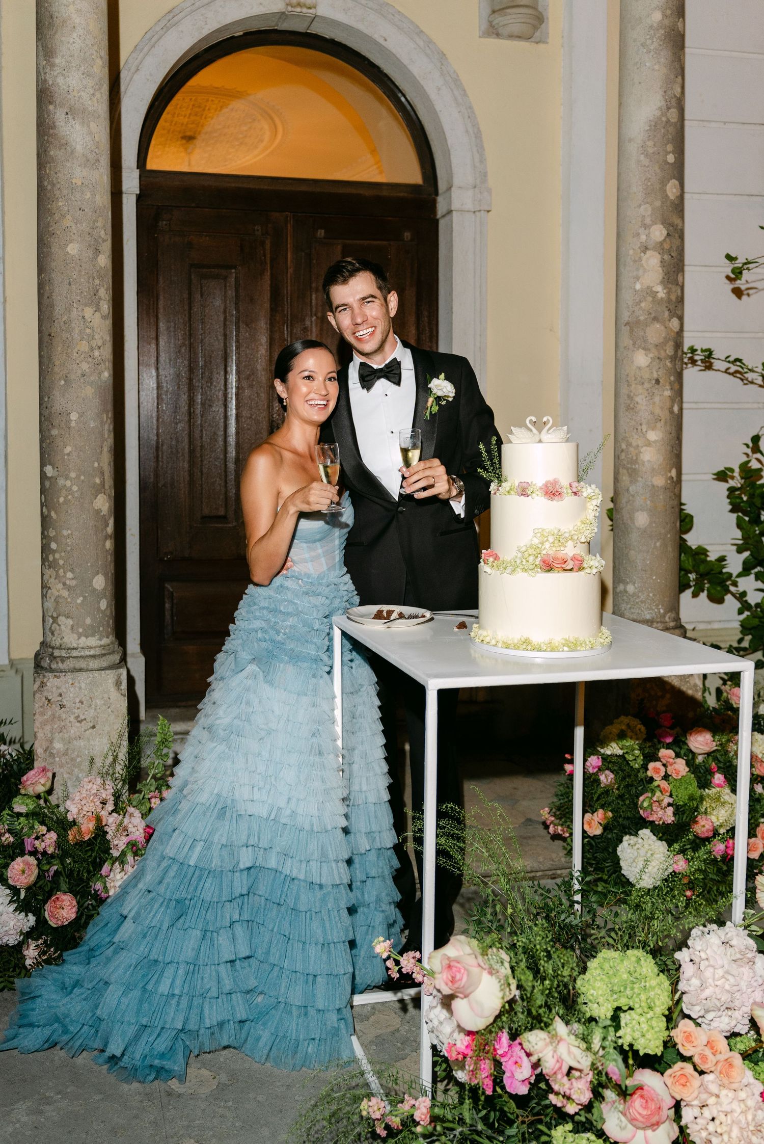 Couple poses with champagne by their elegant floral-adorned wedding cake.