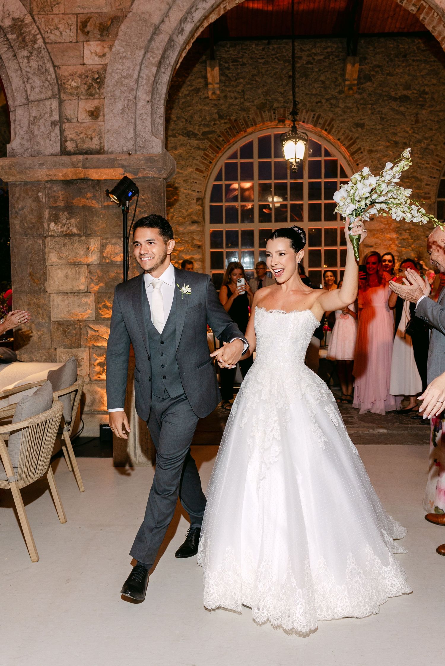A couple dances together at their wedding reception in a rustic stone venue with arched windows.