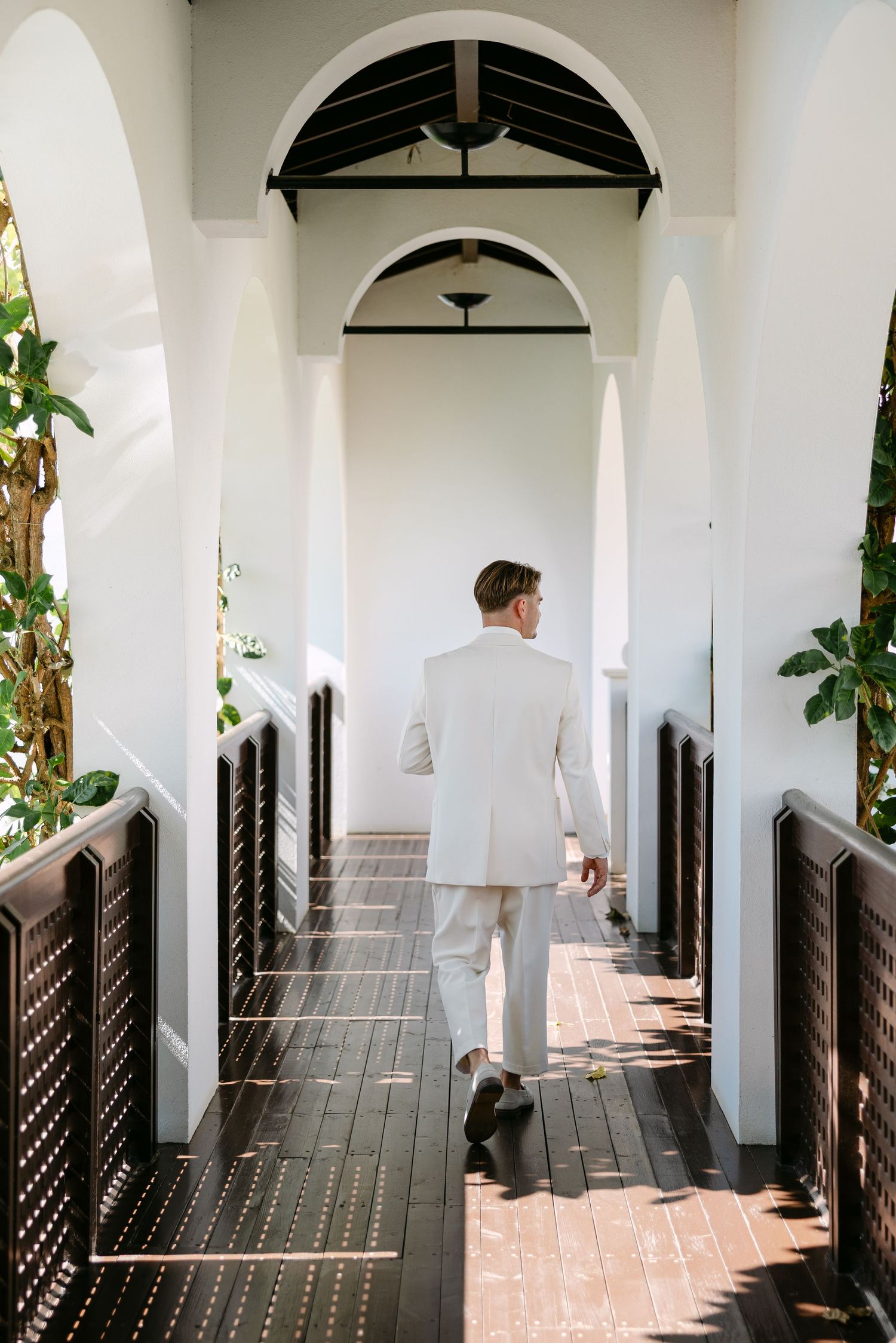 A person in a white suit walks down an elegant arched hallway with white columns and dark wooden shutters.