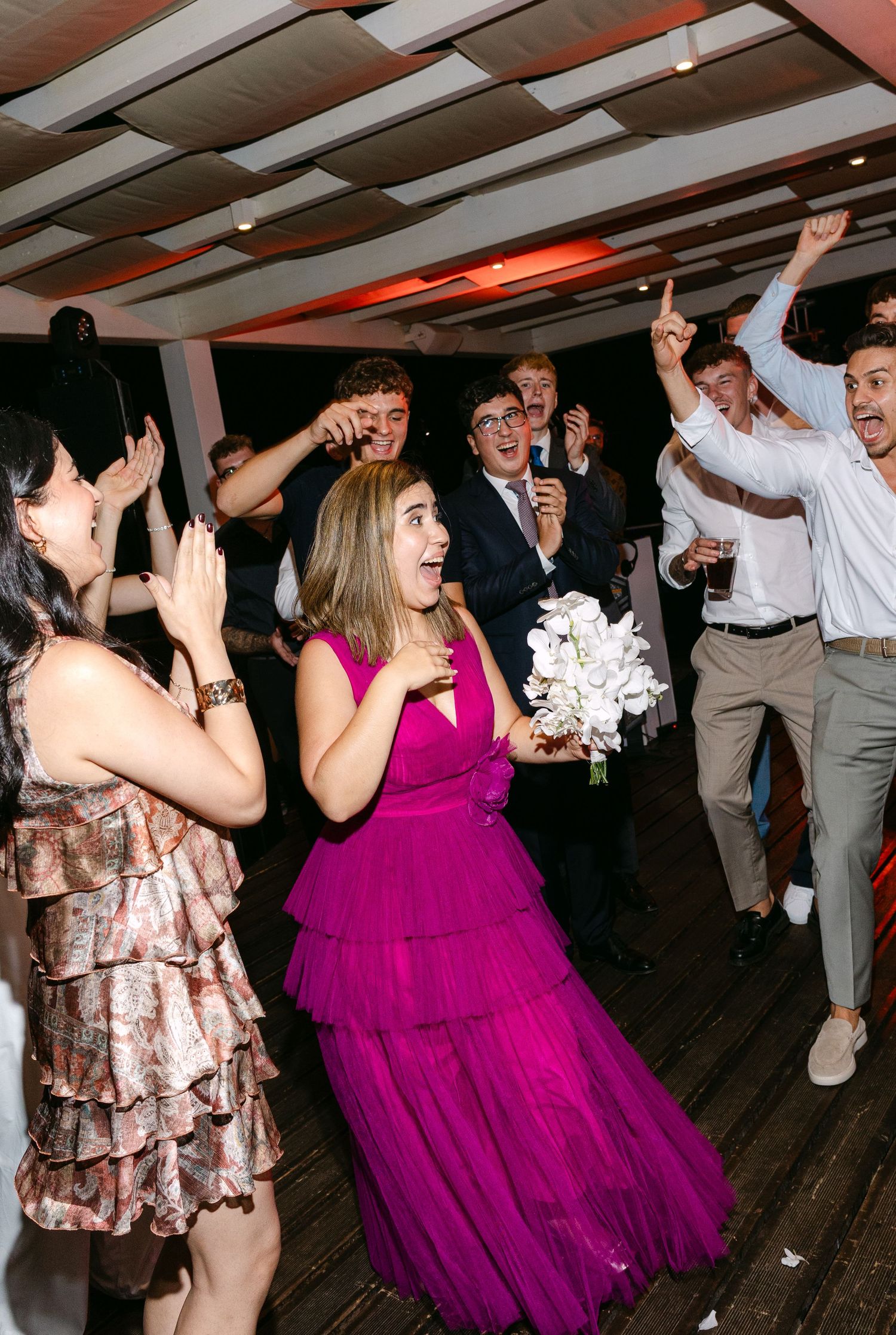 Wedding guests dancing and celebrating with person in bright magenta dress at center of dance floor.