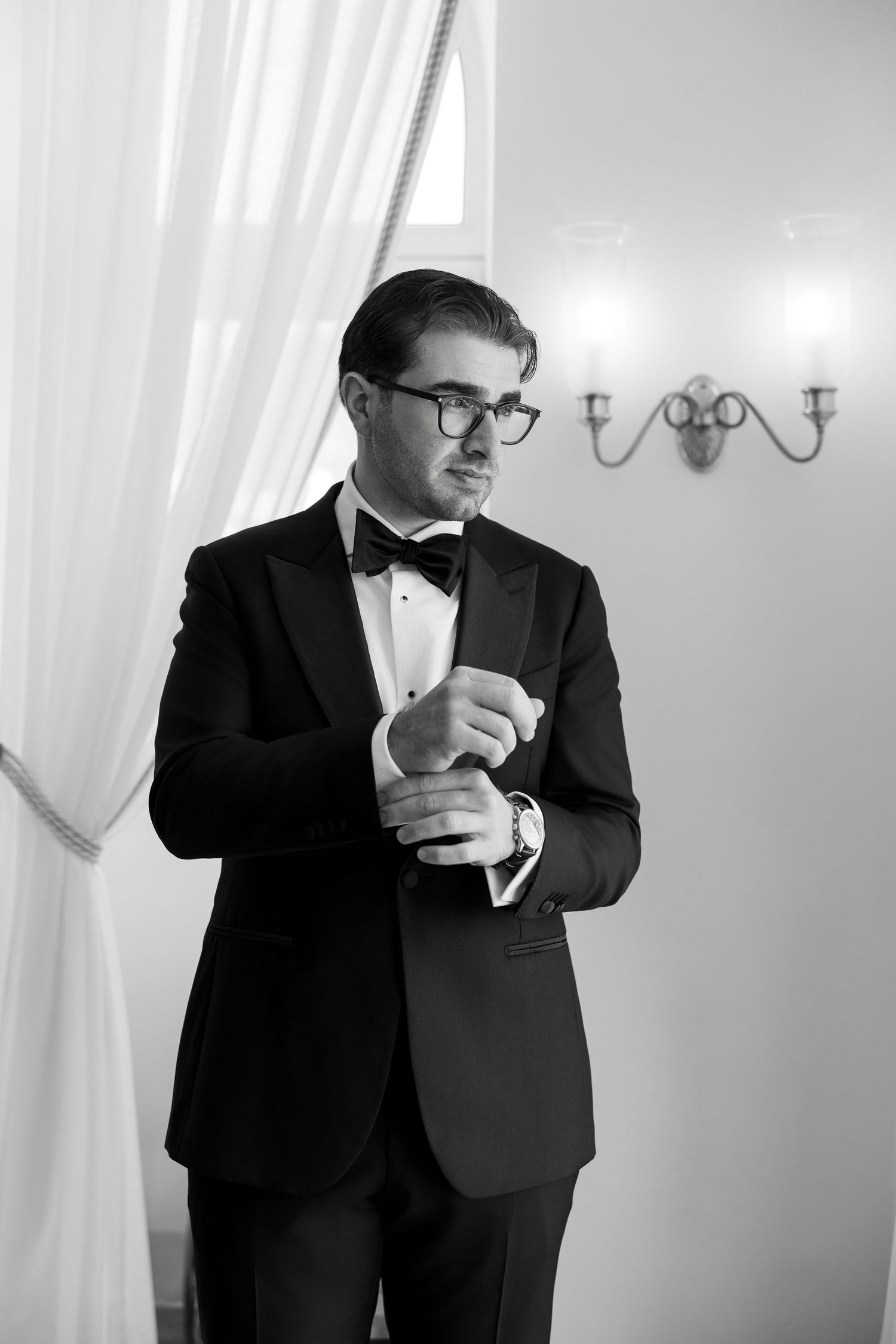 Black and white photo of a groom in a tuxedo and bow tie adjusting his cufflinks before his wedding ceremony.