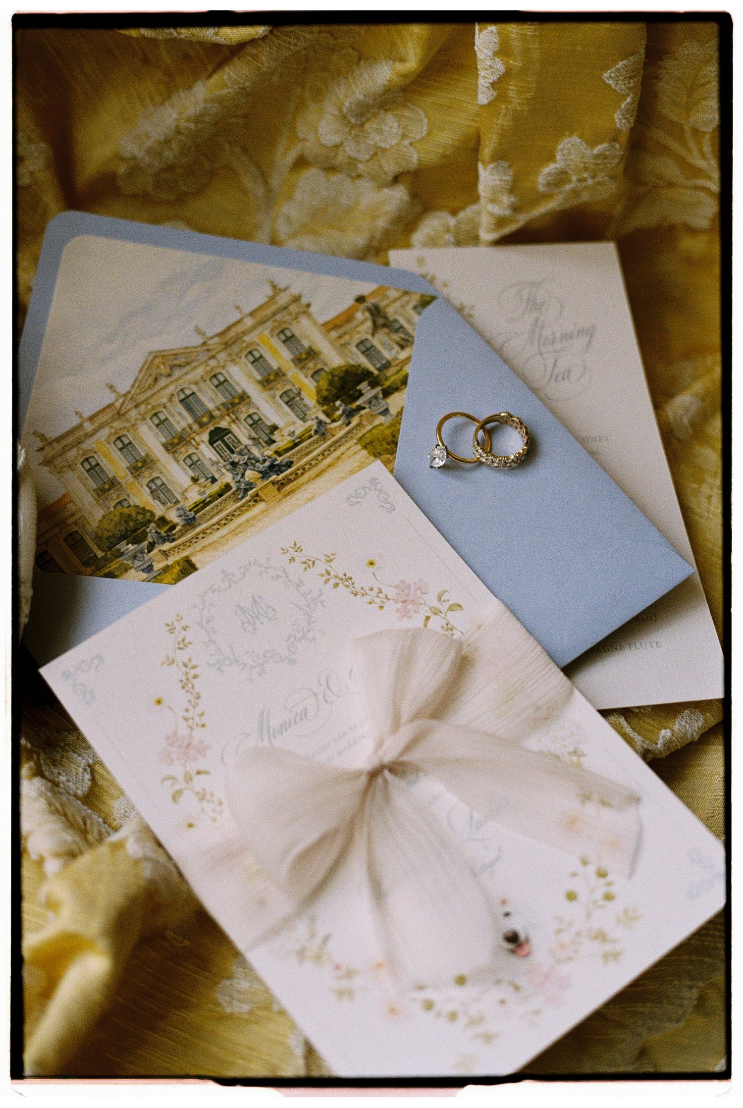 Elegant wedding stationery flat lay with floral invitation, blue envelope, venue illustration, and diamond rings on gold fabric.