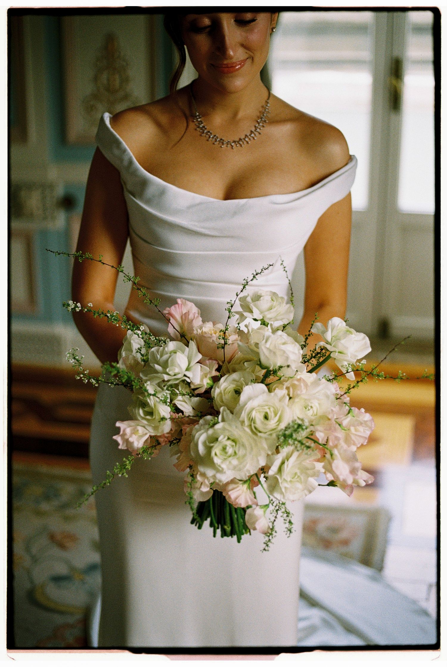 Bride in off-shoulder white gown holding lush bouquet of white and blush roses with delicate greenery.