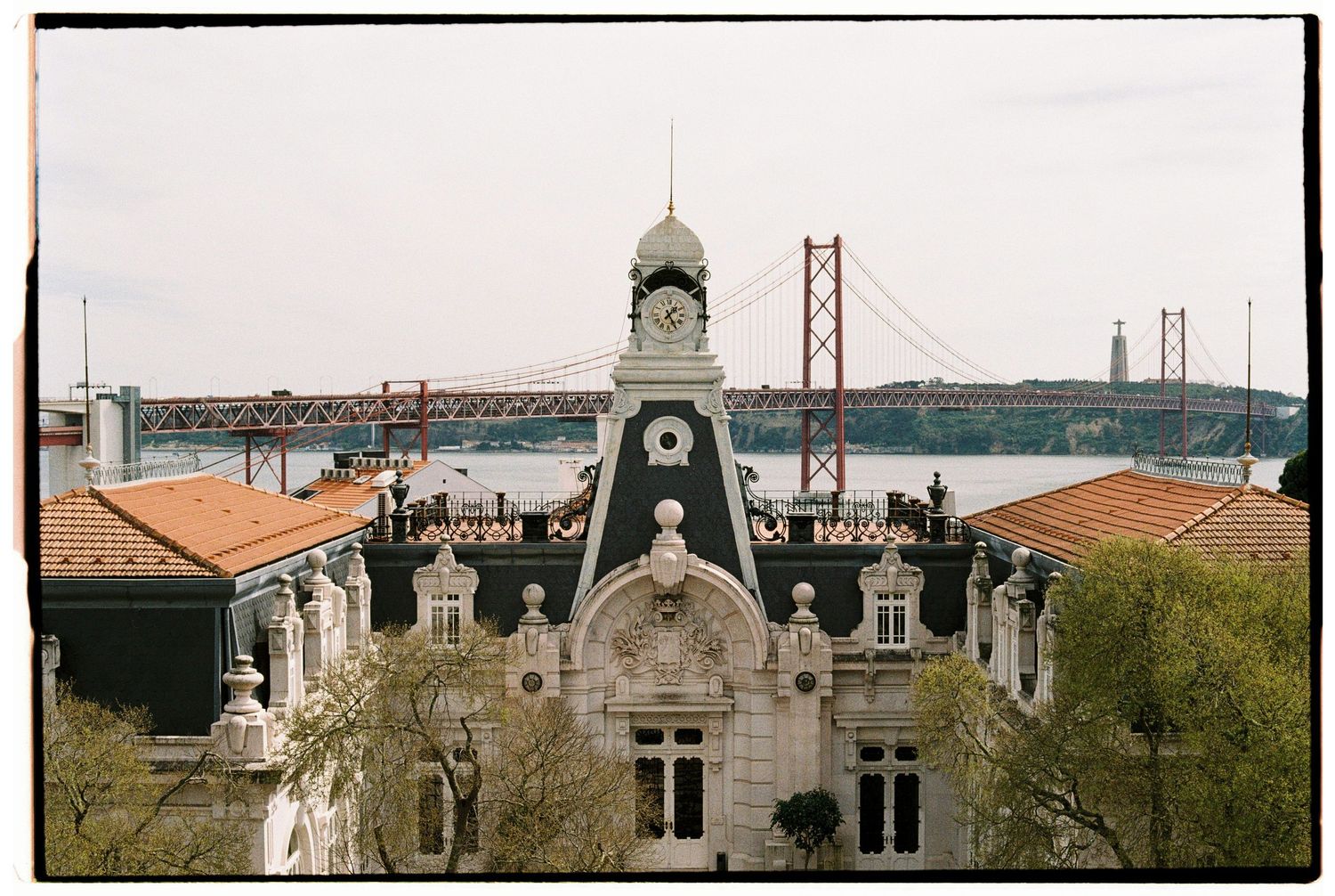 Historic building with ornate clock tower overlooking the Tagus River and the 25 de Abril Bridge in Lisbon, Portugal.