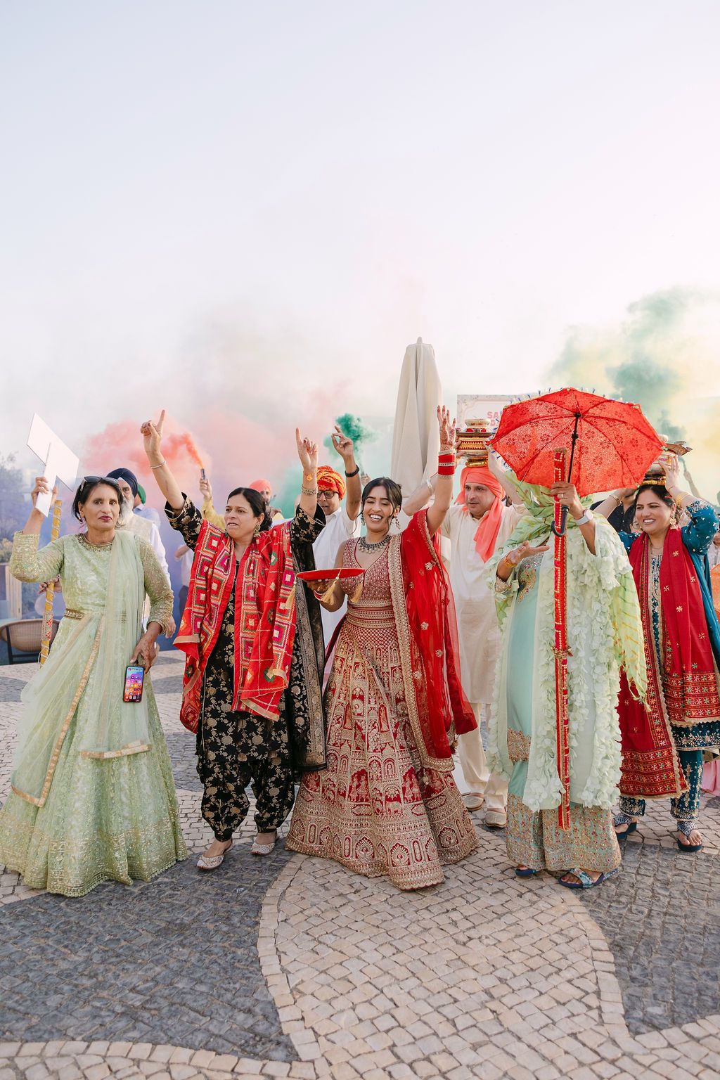 Wedding party celebrates in colorful traditional Indian attire with red umbrellas and vibrant lehengas on cobblestone path.