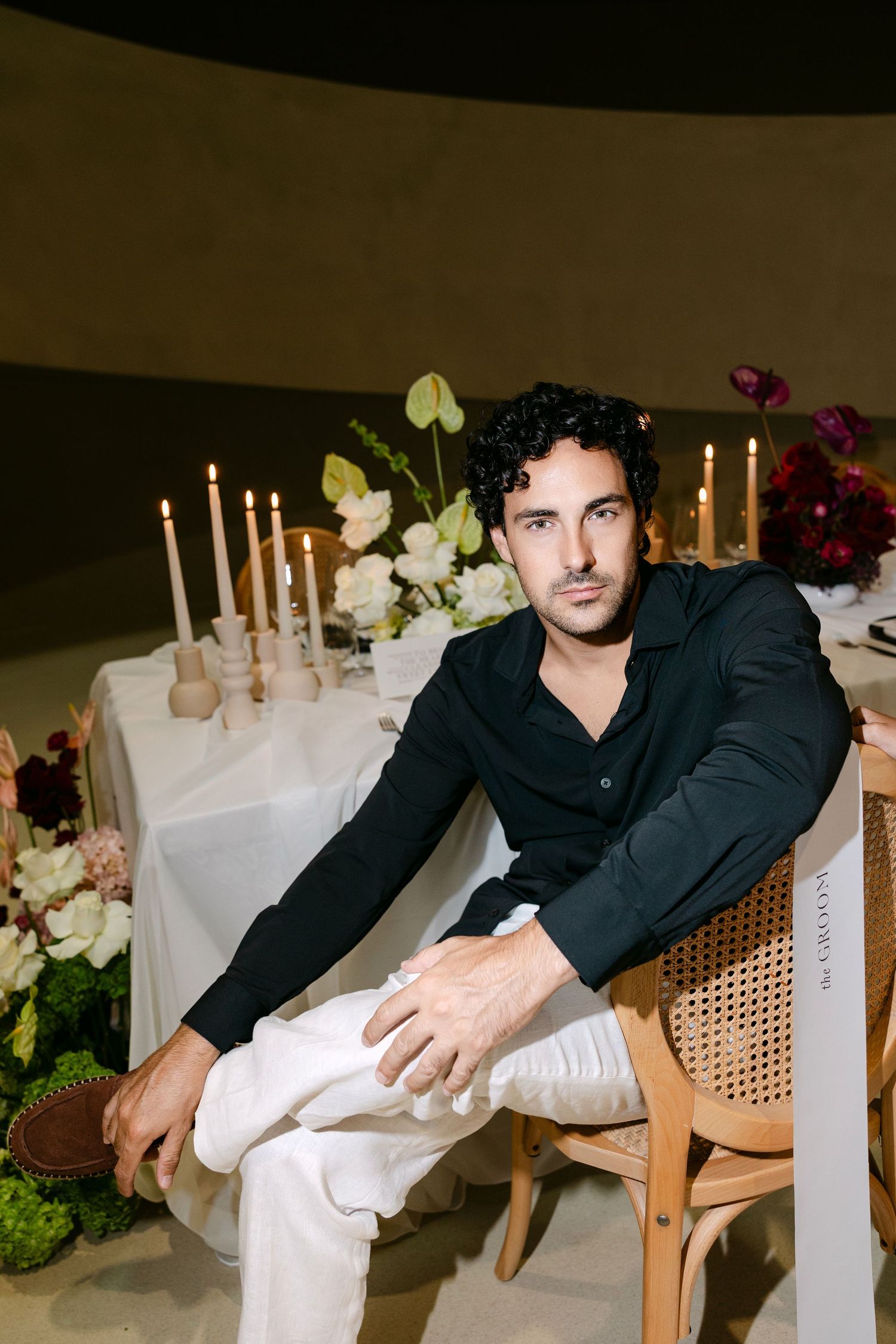Stylish man in black shirt and white pants sits relaxed at a candlelit floral wedding reception table.