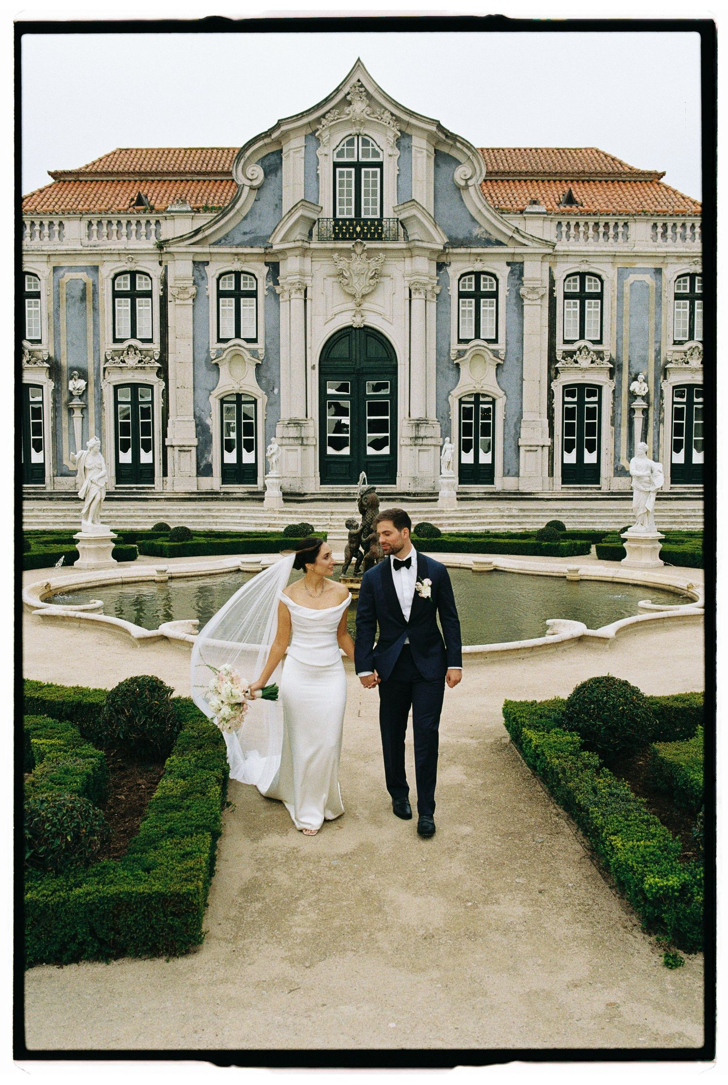 Bride and groom walk hand-in-hand through formal gardens before an ornate Baroque palace in Portugal.