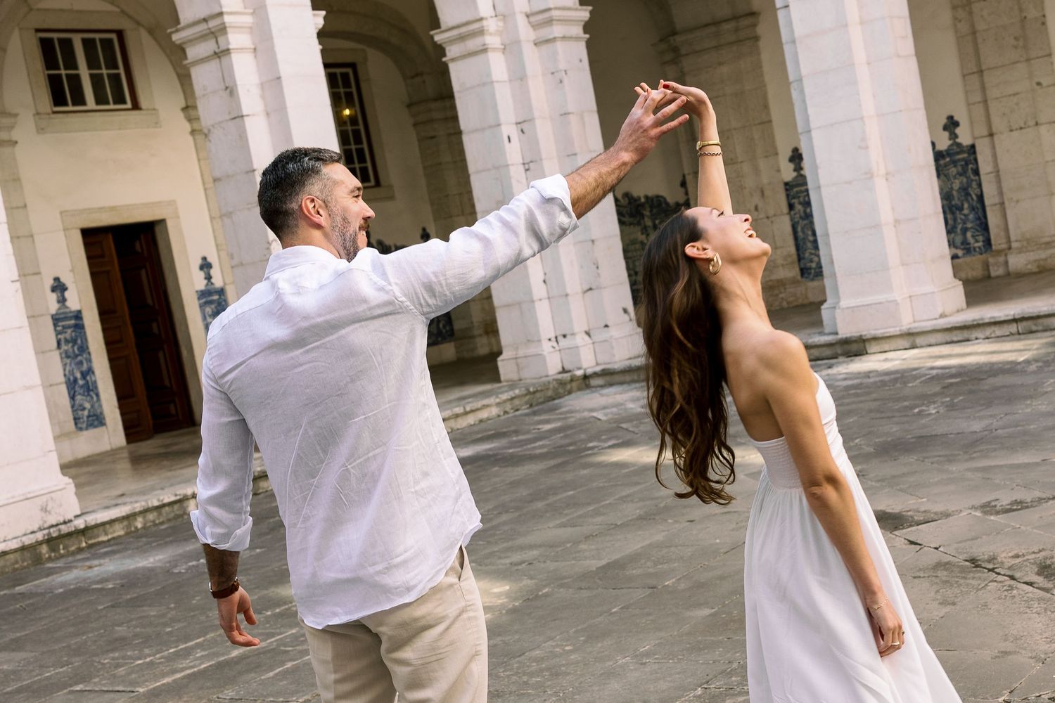 Couple dancing outdoors in front of a historic building, man in white shirt spinning woman in elegant white dress.