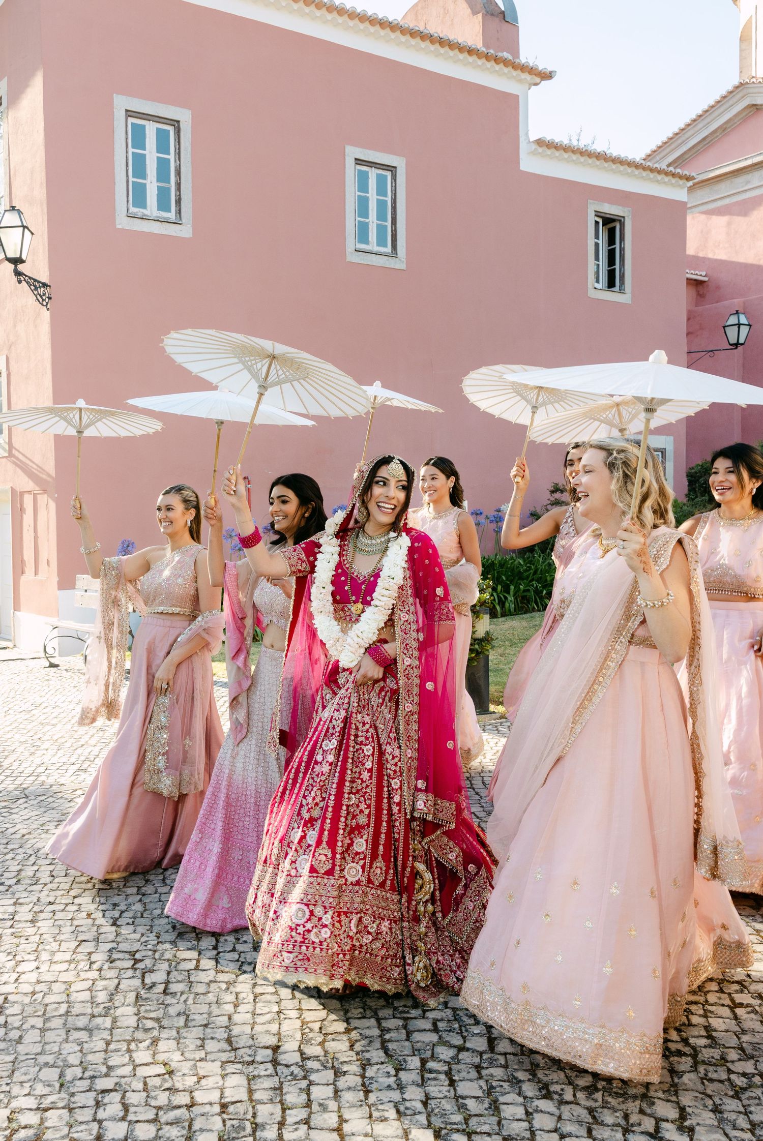 Bridal party in pink and red lehengas posing with parasols on cobblestone street.