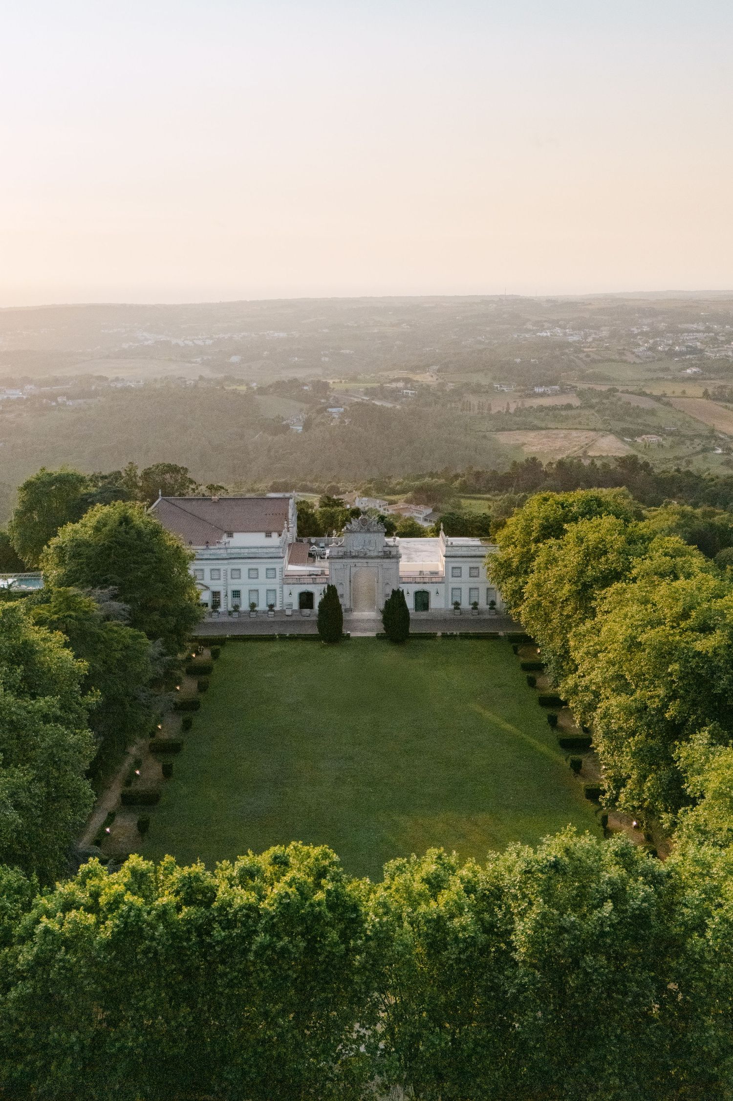 Aerial view of an elegant white mansion with manicured gardens and sprawling countryside views.