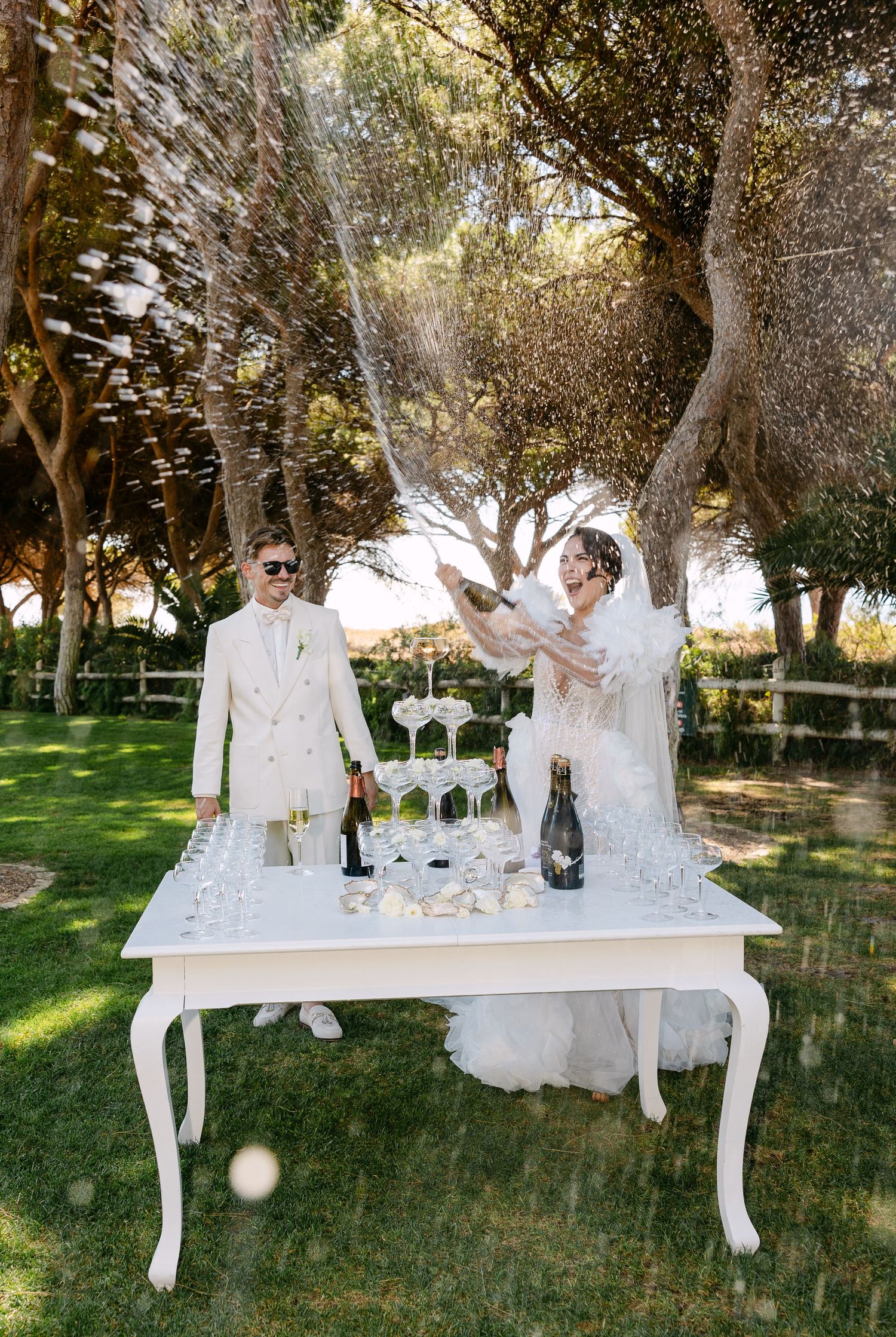 A couple in white attire celebrates by spraying champagne at an outdoor wedding reception.