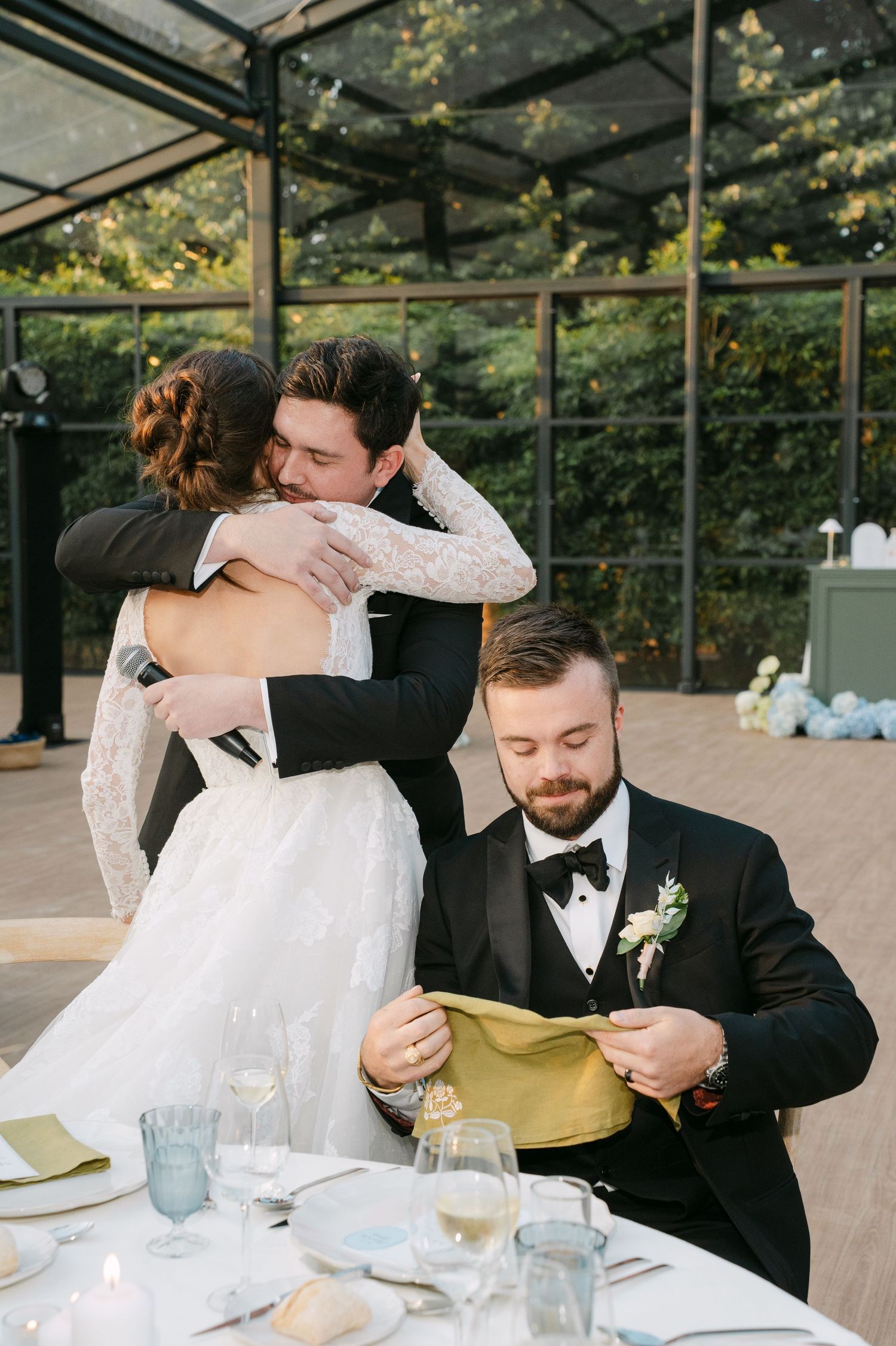 Wedding reception moment in a glass-enclosed venue with guests at dining tables.