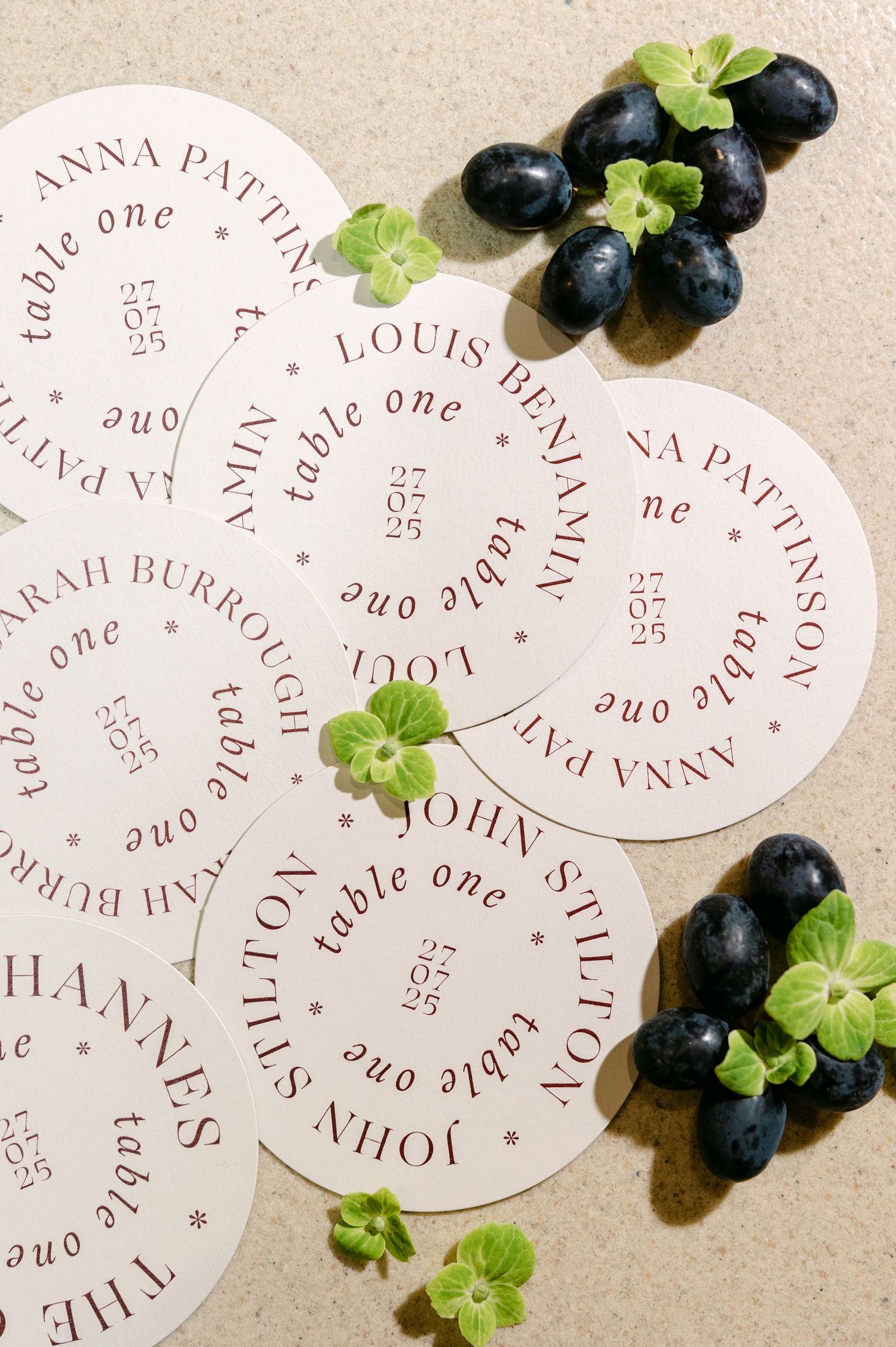 Circular wedding place cards with burgundy text arranged with fresh blueberries and green herbs.