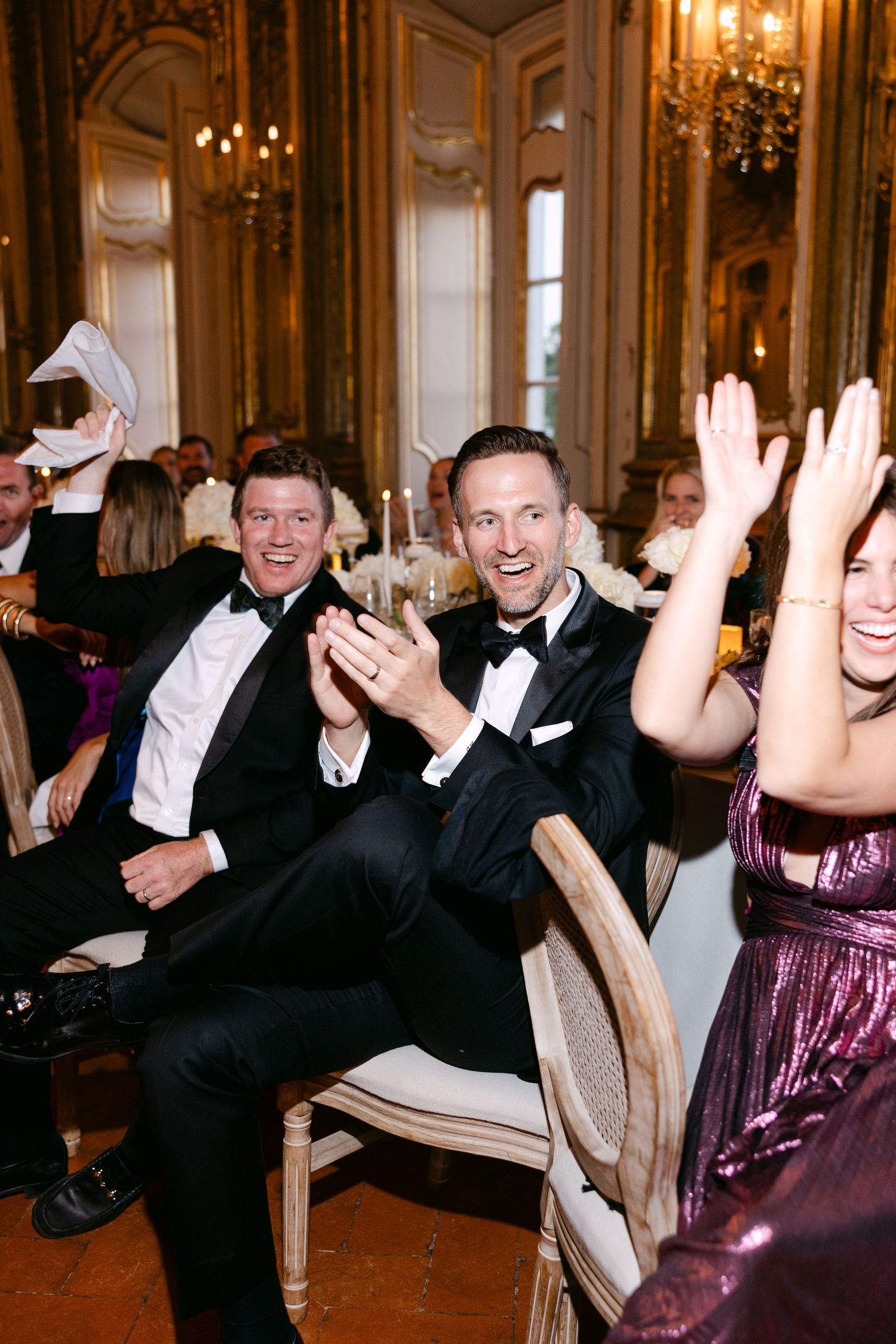 Wedding guests laugh and celebrate in opulent ballroom with chandeliers.