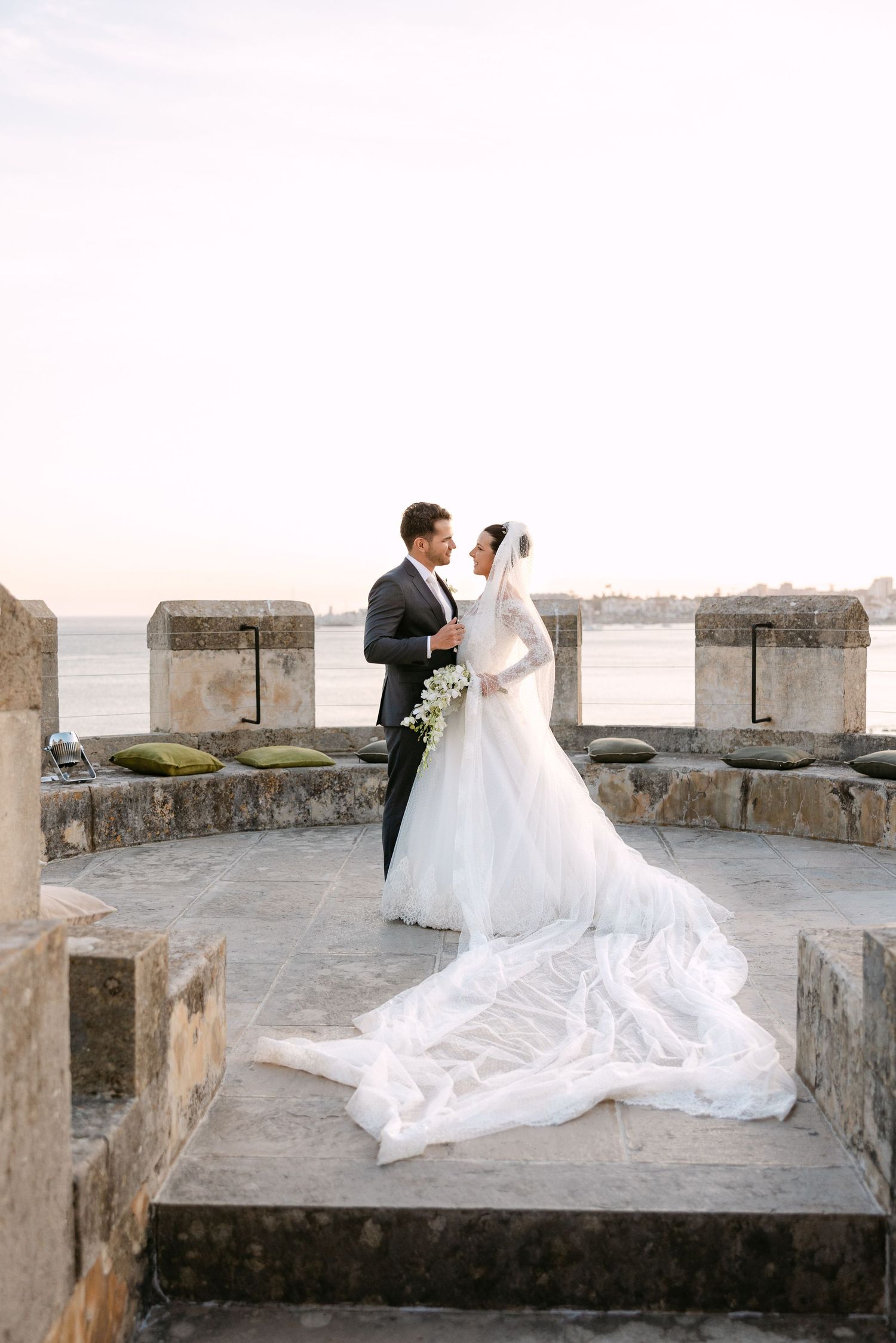 Wedding couple in elegant attire posing along stone balcony with flowing white dress train.
