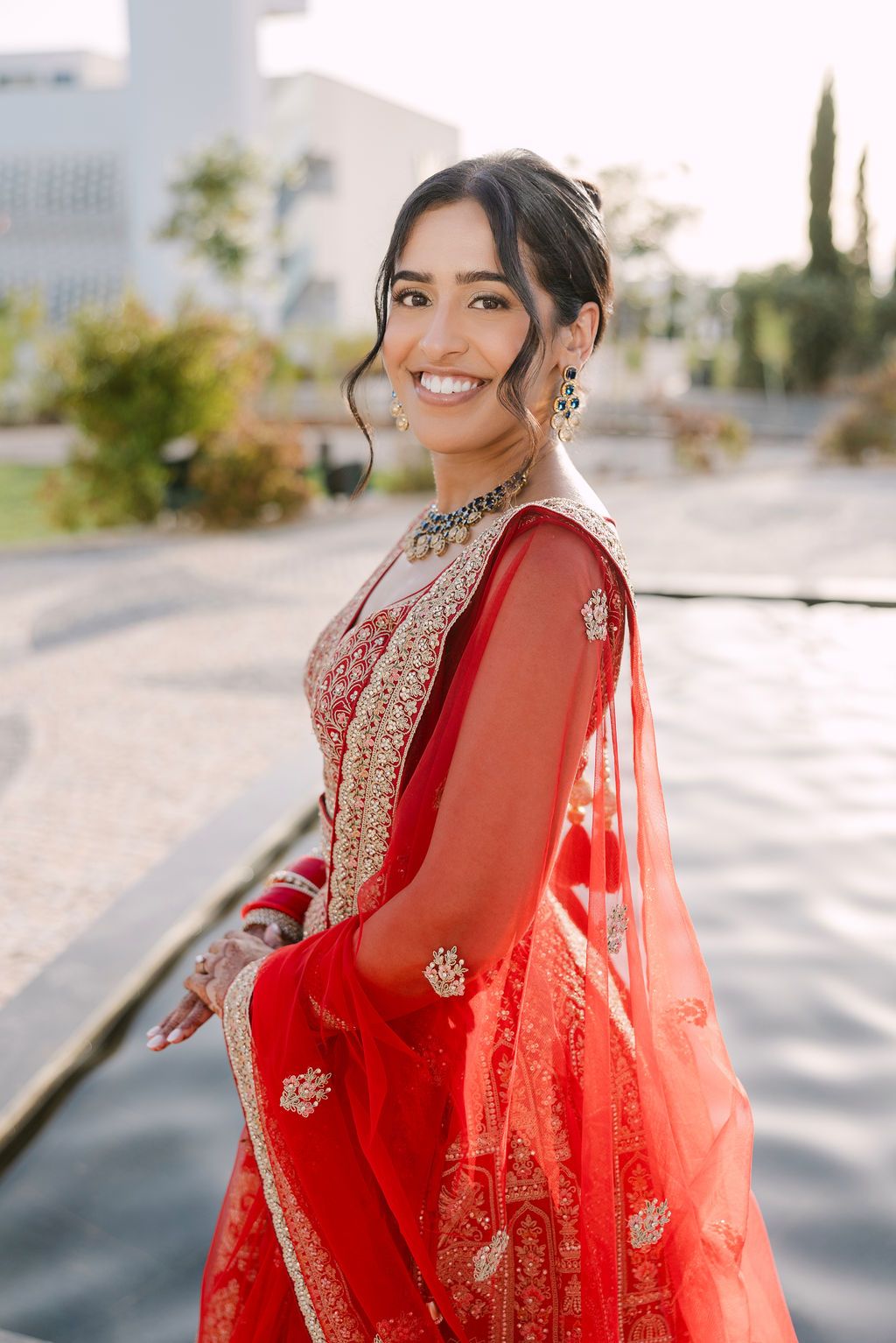 Elegant bridal portraits featuring red and gold embroidered lehenga with flowing dupatta in sunlight.