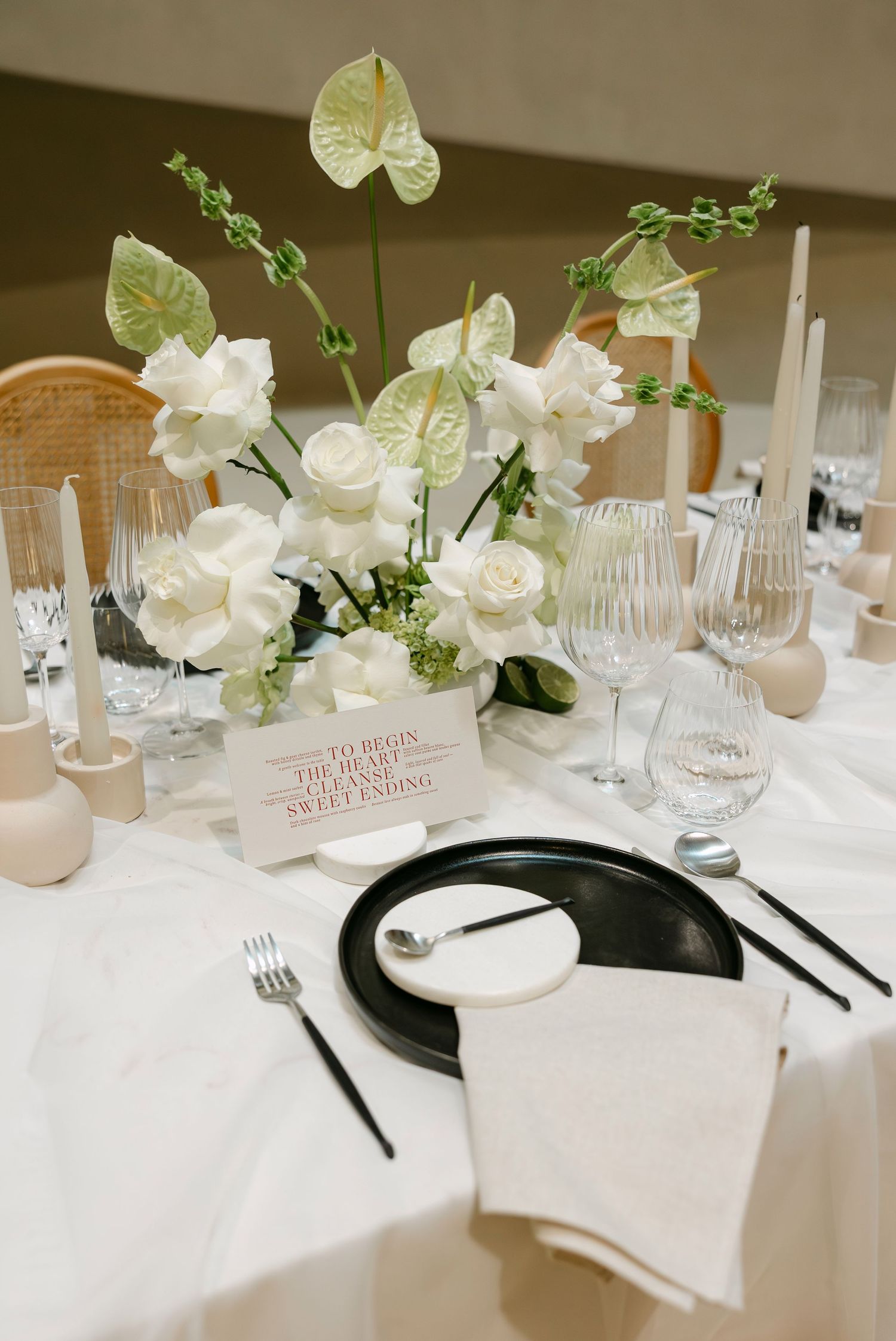 Elegant wedding table setting with white roses, anthurium centerpiece, black plates, taper candles, and crystal glassware on white linen.